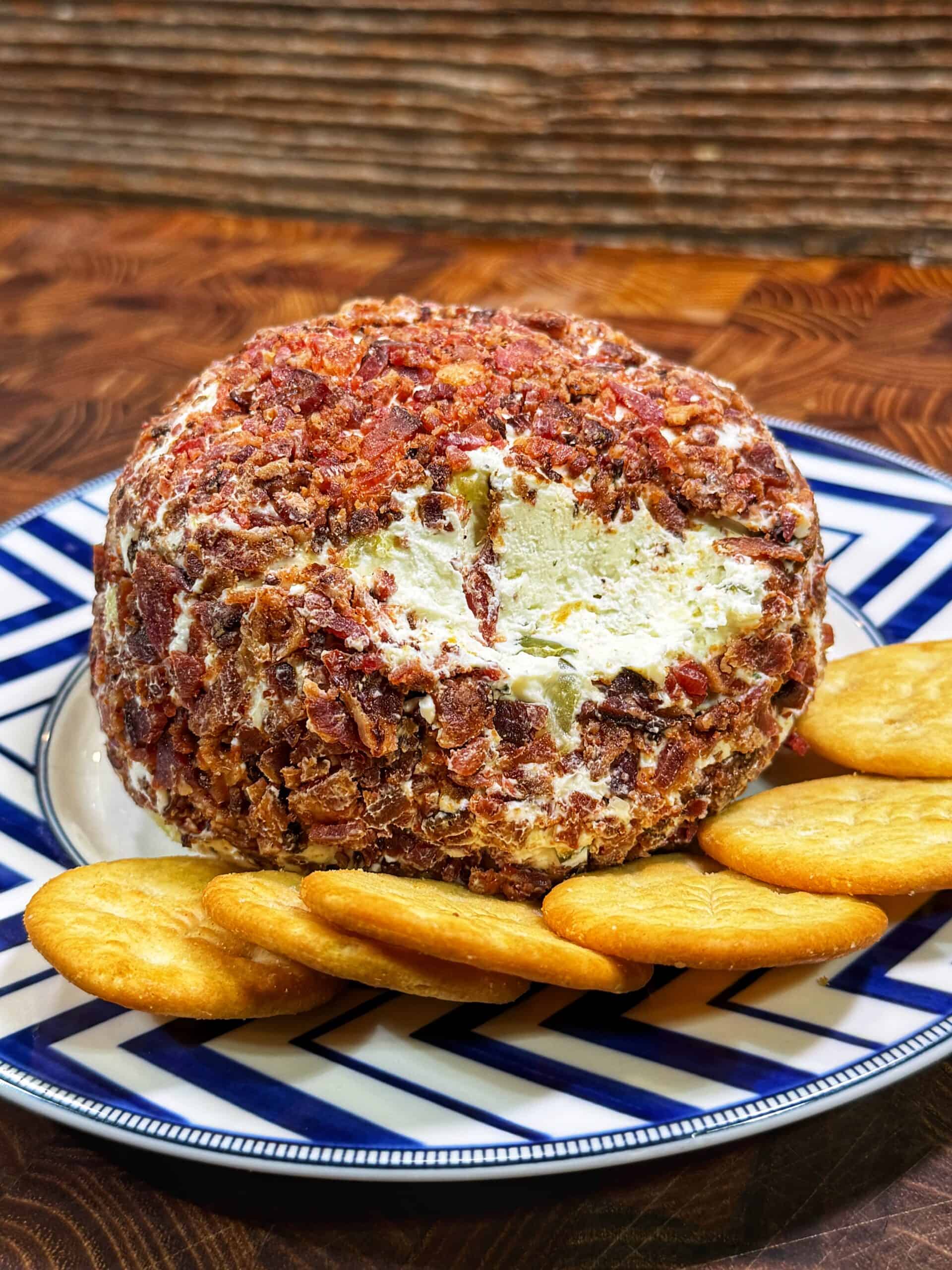 A round cheese ball coated with chopped bacon is served on a patterned plate with several round crackers arranged around it. A portion of the cheese ball has been scooped out, revealing the creamy interior.