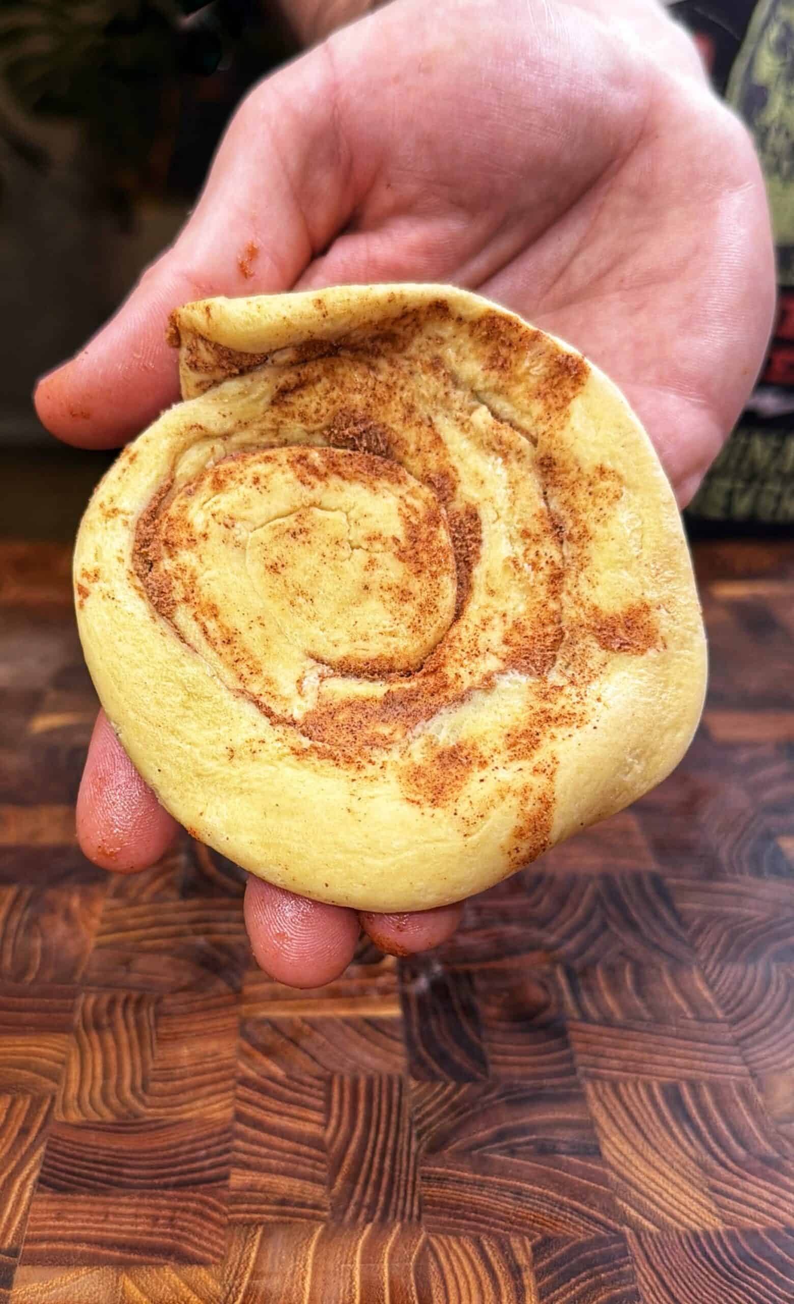 A hand holding an unbaked cinnamon roll dough spiral over a wooden surface, showing swirls of cinnamon filling.