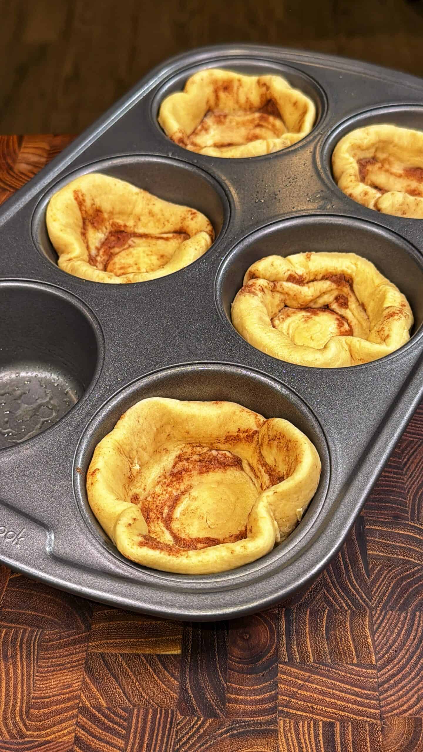A muffin tin containing five baked, golden brown cinnamon swirl pastry cups, resting on a wooden surface. One muffin slot is empty, showing a bit of nonstick spray residue.