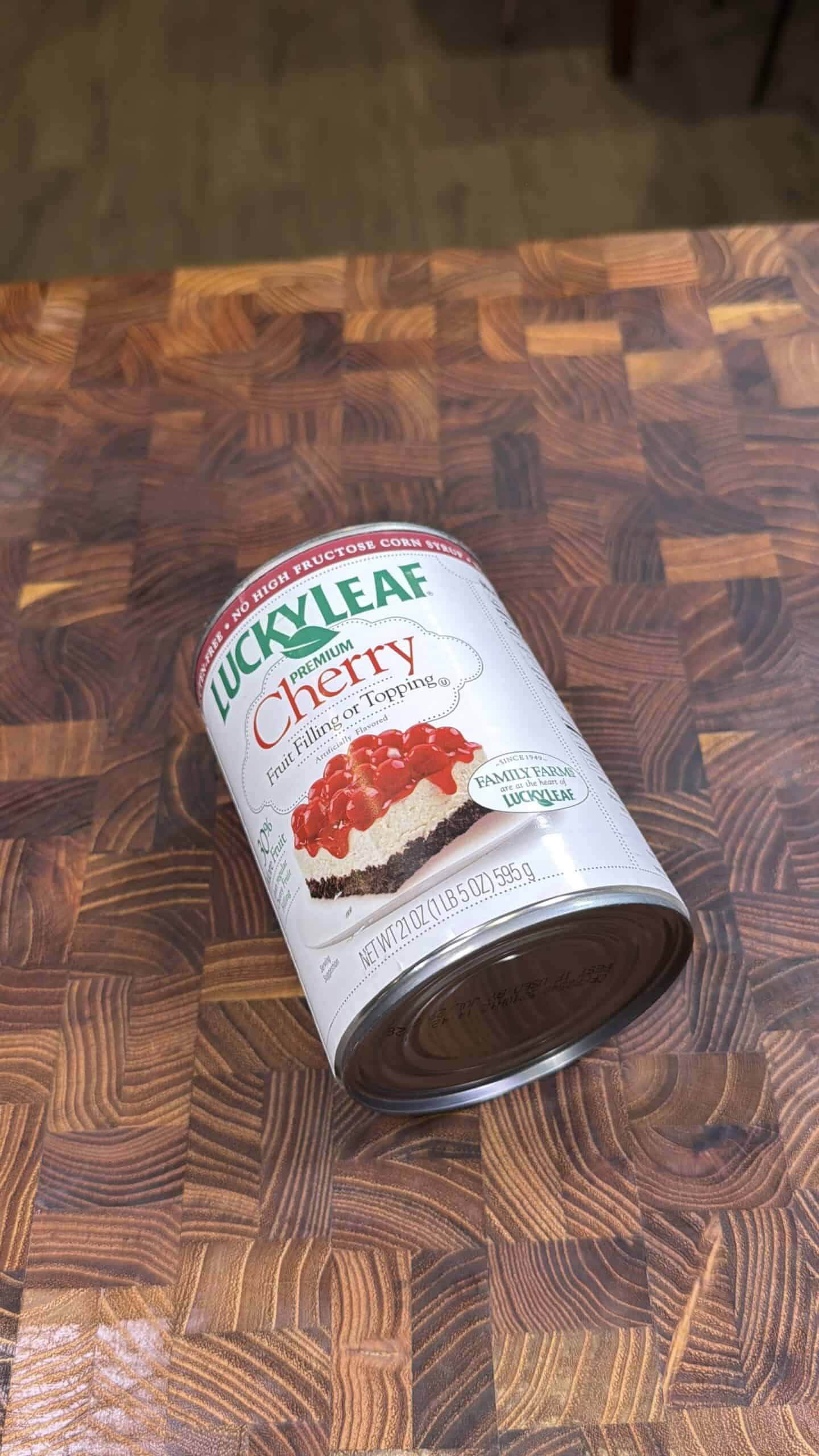 A can of Lucky Leaf Premium Cherry fruit filling and topping is lying on its side on a wooden checkered cutting board. The can features an image of cherry-topped dessert on the label.