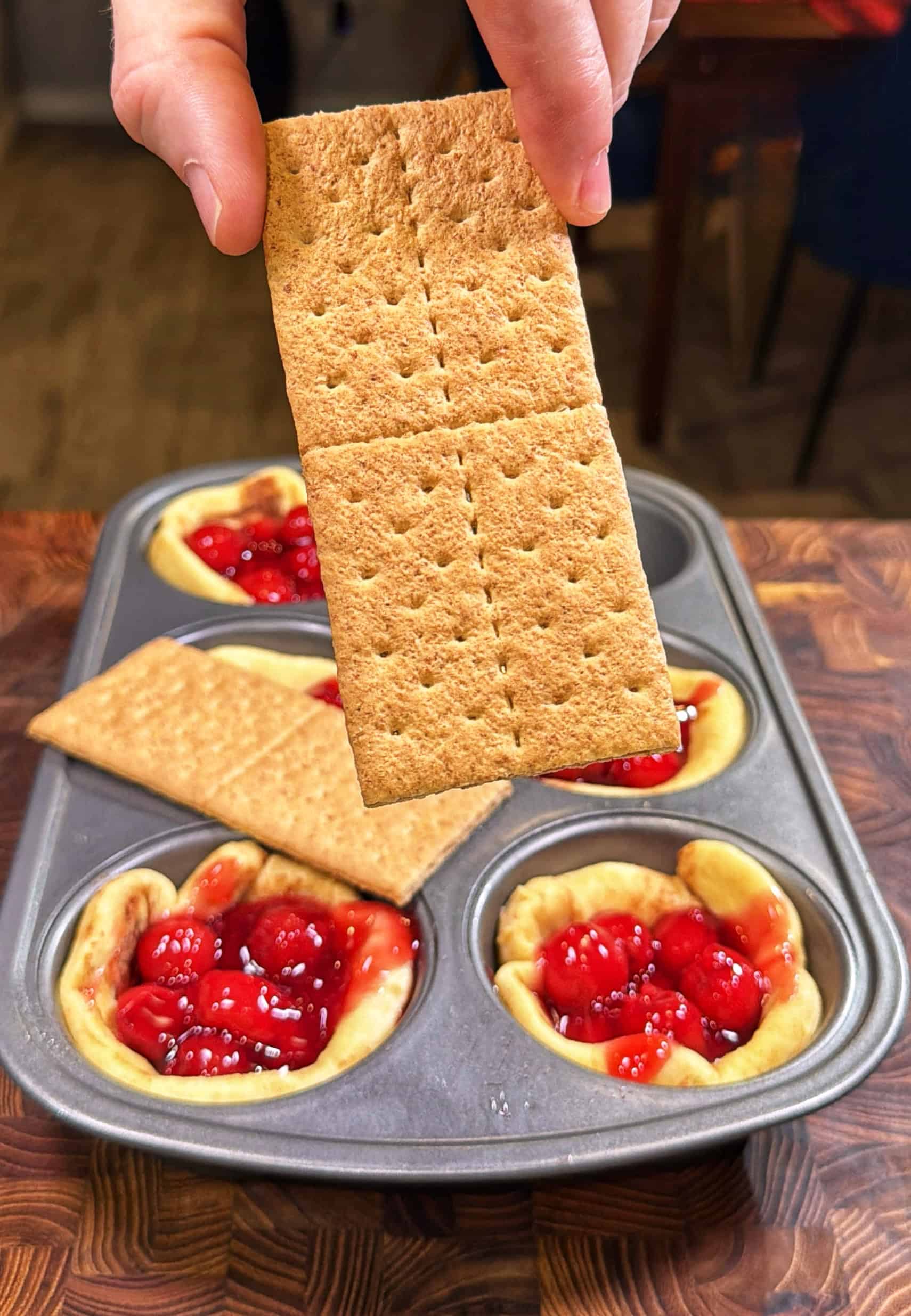 A hand holds a rectangular graham cracker over a muffin tin filled with mini cherry pies, some topped with graham crackers and others showing cherry filling.