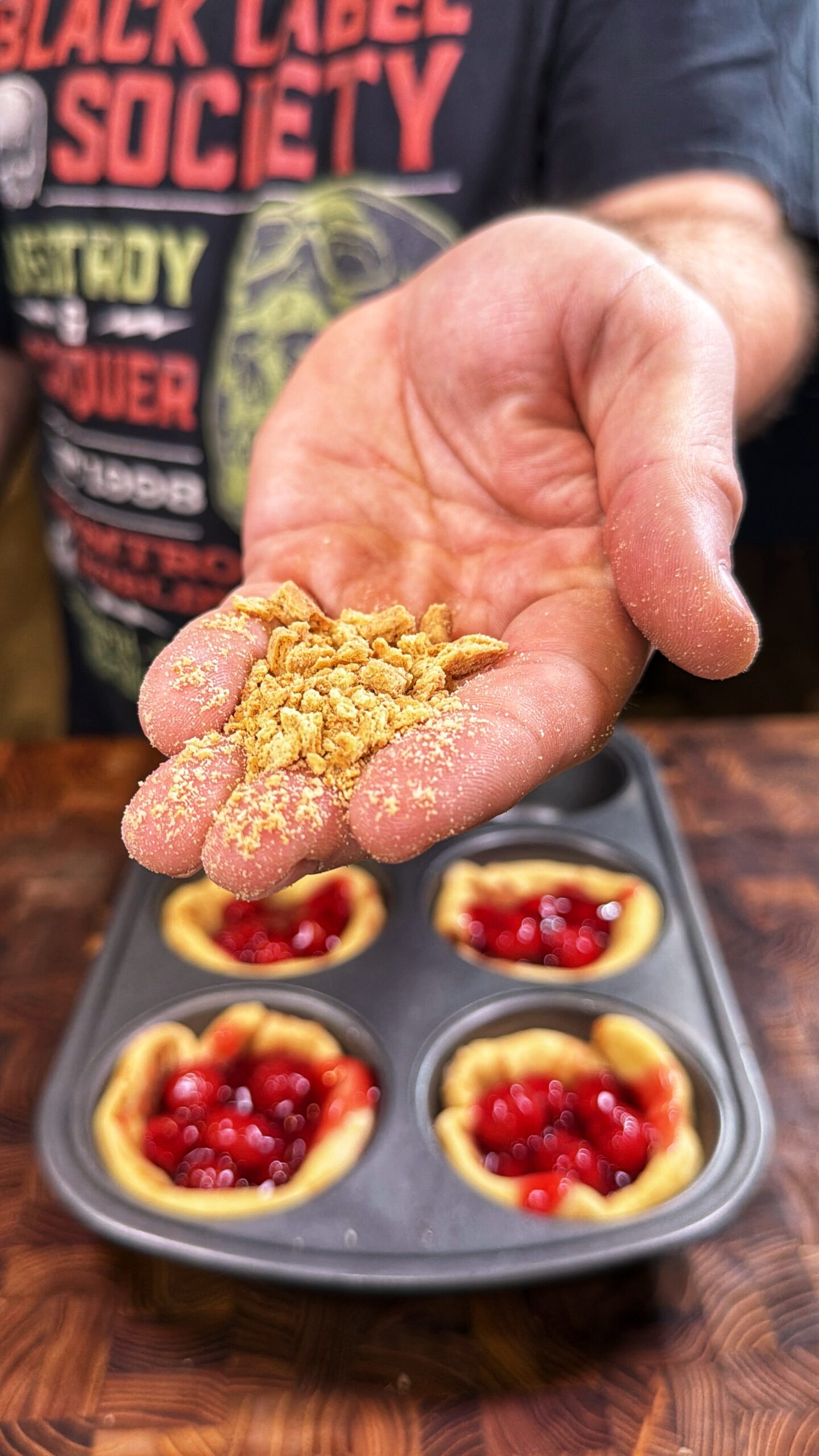 A hand holds a handful of crumbled topping above a muffin tin filled with unbaked cherry tarts. The person is wearing a Black Label Society t-shirt.
