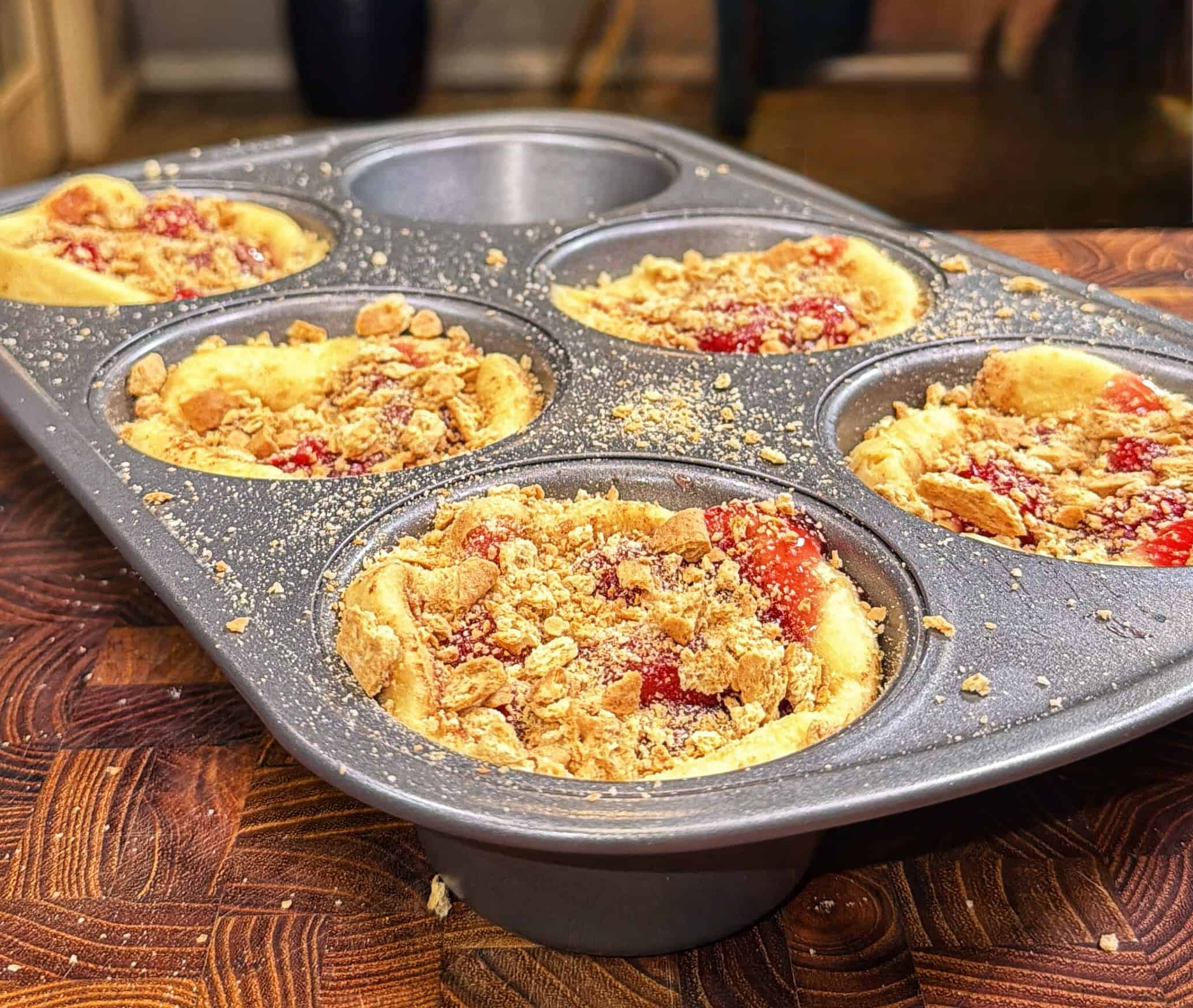 A muffin tin filled with five mini tarts, topped with a crumbly streusel and red fruit filling, sits on a wooden surface in a kitchen setting.