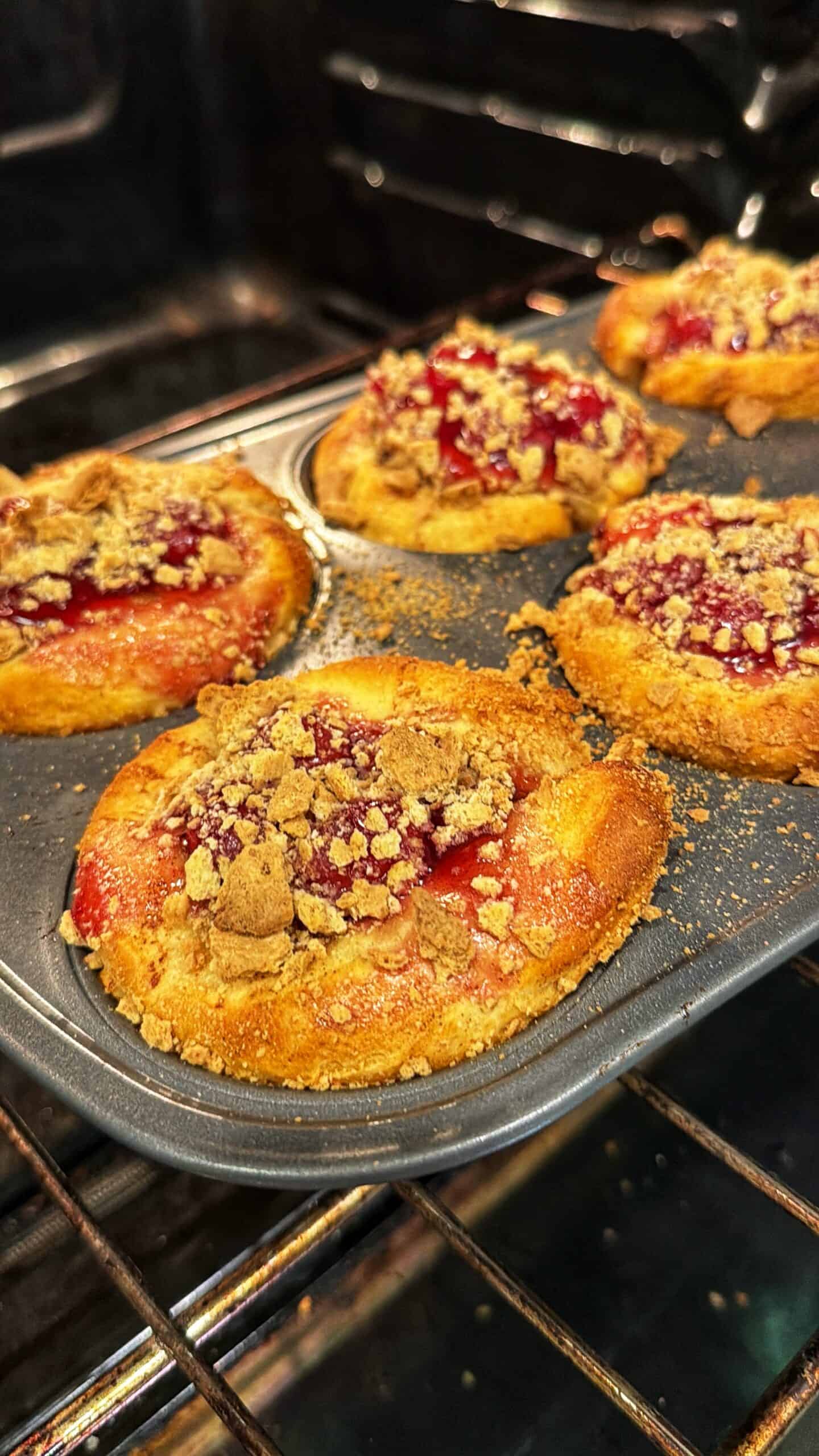Six freshly baked pastries with a crumb topping and red fruit filling are in a muffin tin inside an oven. The golden-brown crust and bubbling filling suggest they are nearly ready to be taken out.
