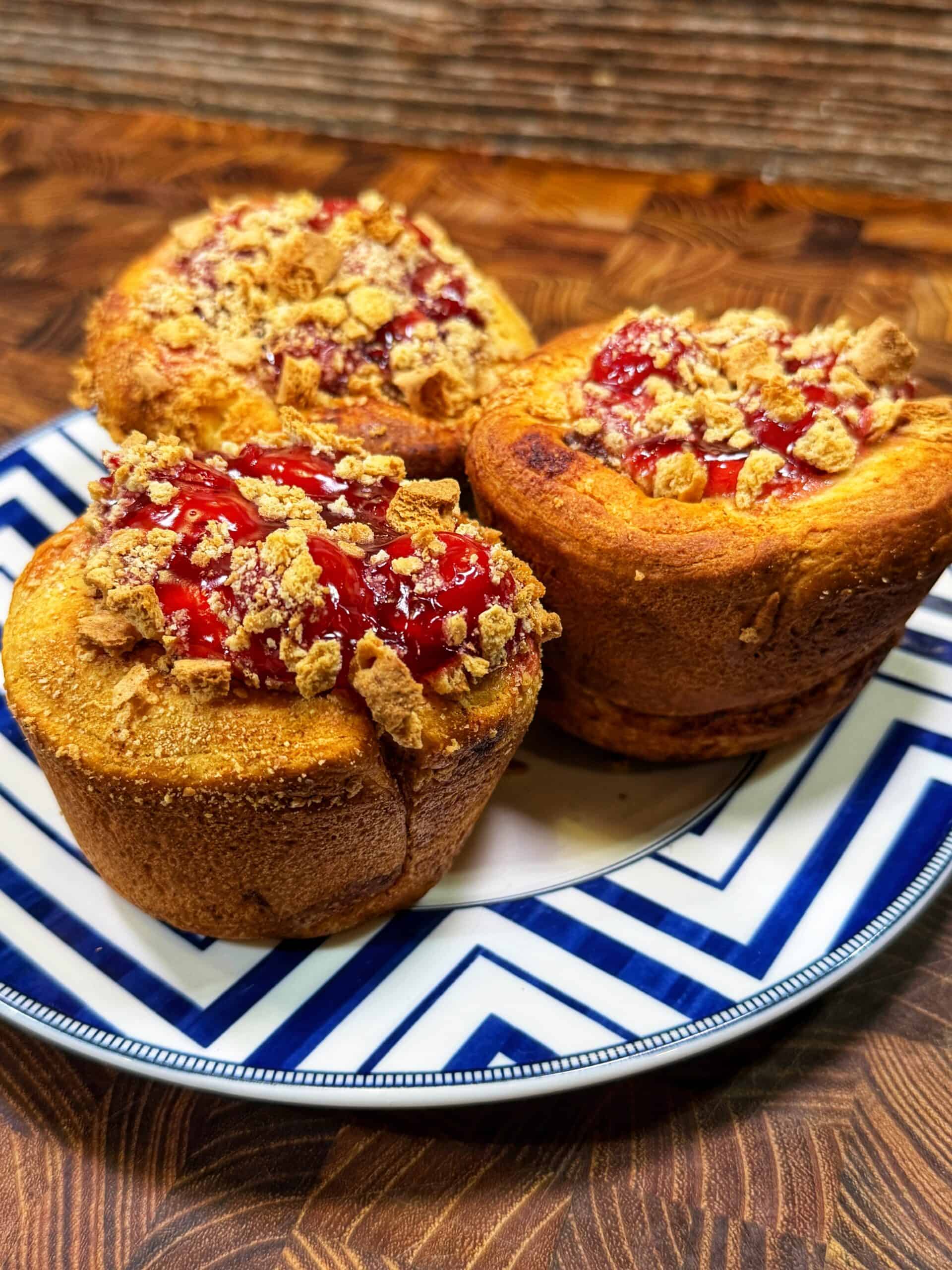 Three golden brown muffins topped with crushed crumbs and a red fruit glaze sit on a blue and white zigzag-patterned plate, placed on a wooden surface.