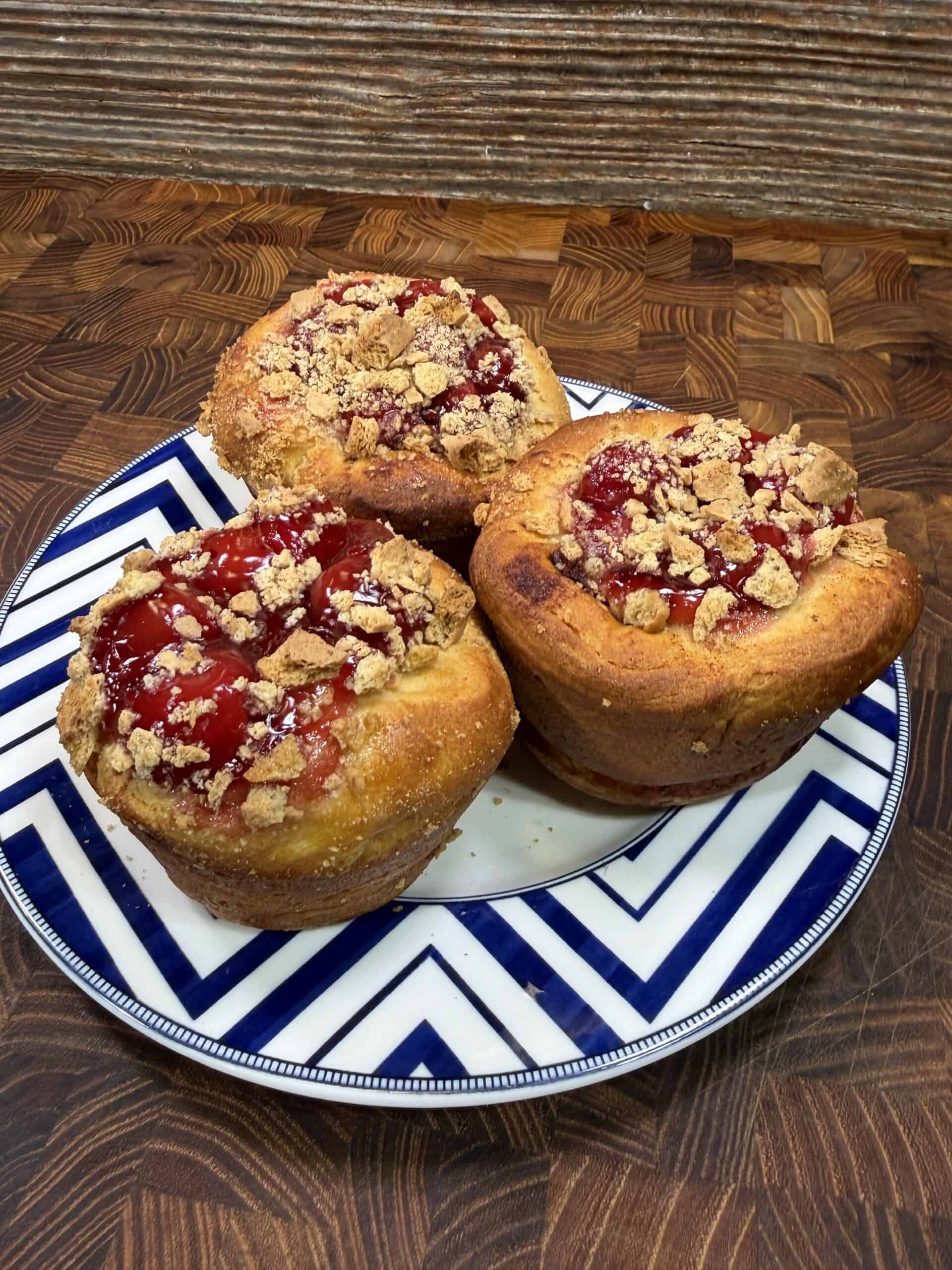 Three large muffin-sized pastries topped with cherry filling and crumbly streusel sit on a white plate with blue zigzag patterns. The plate is on a wooden surface with a textured brown background.