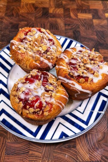 Three pastries with a fruit filling, streusel topping, and white icing drizzle are arranged on a blue-and-white zigzag patterned plate atop a wooden surface.
