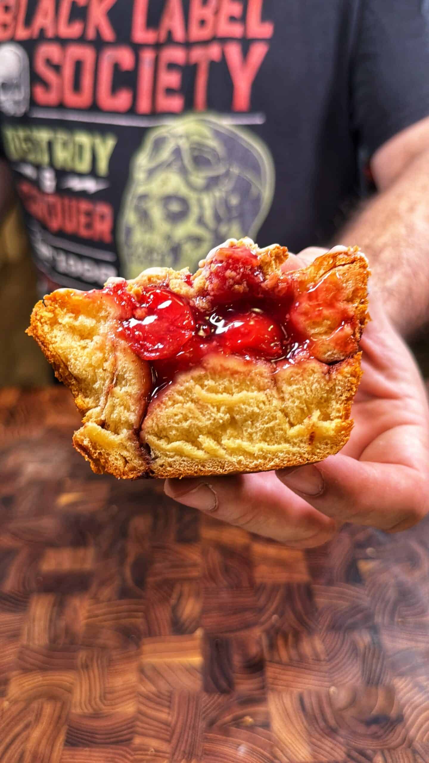 A person holding a thick slice of bread pudding topped with red fruit sauce, with a person wearing a Black Label Society shirt in the background. Recipe is for cherry stuffed cinnamon roll muffins