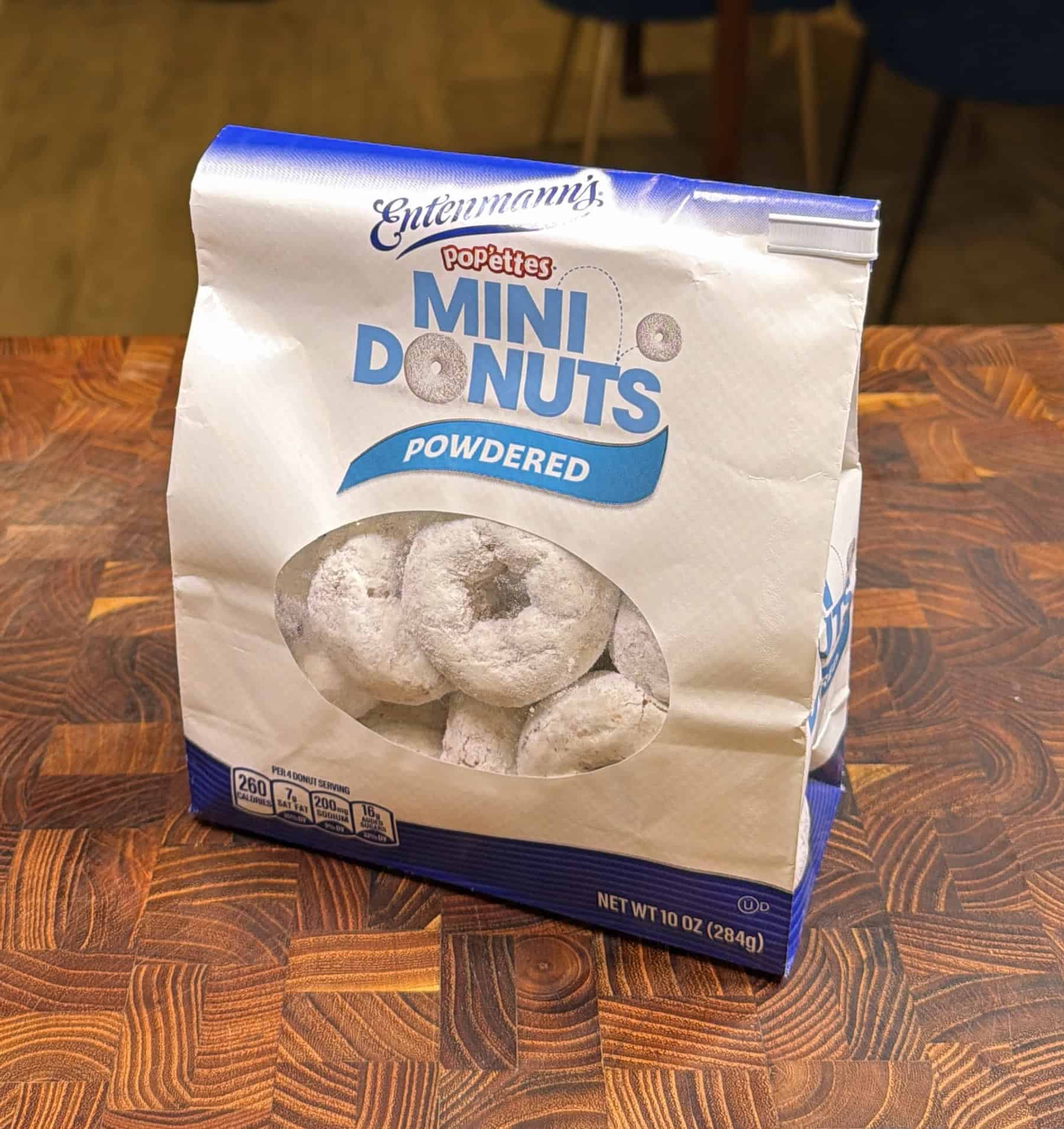 A bag of Entenmann’s Pop‘ettes Mini Donuts Powdered sits on a wooden table, showing round, white powdered donuts visible through a clear window on the front of the packaging.
