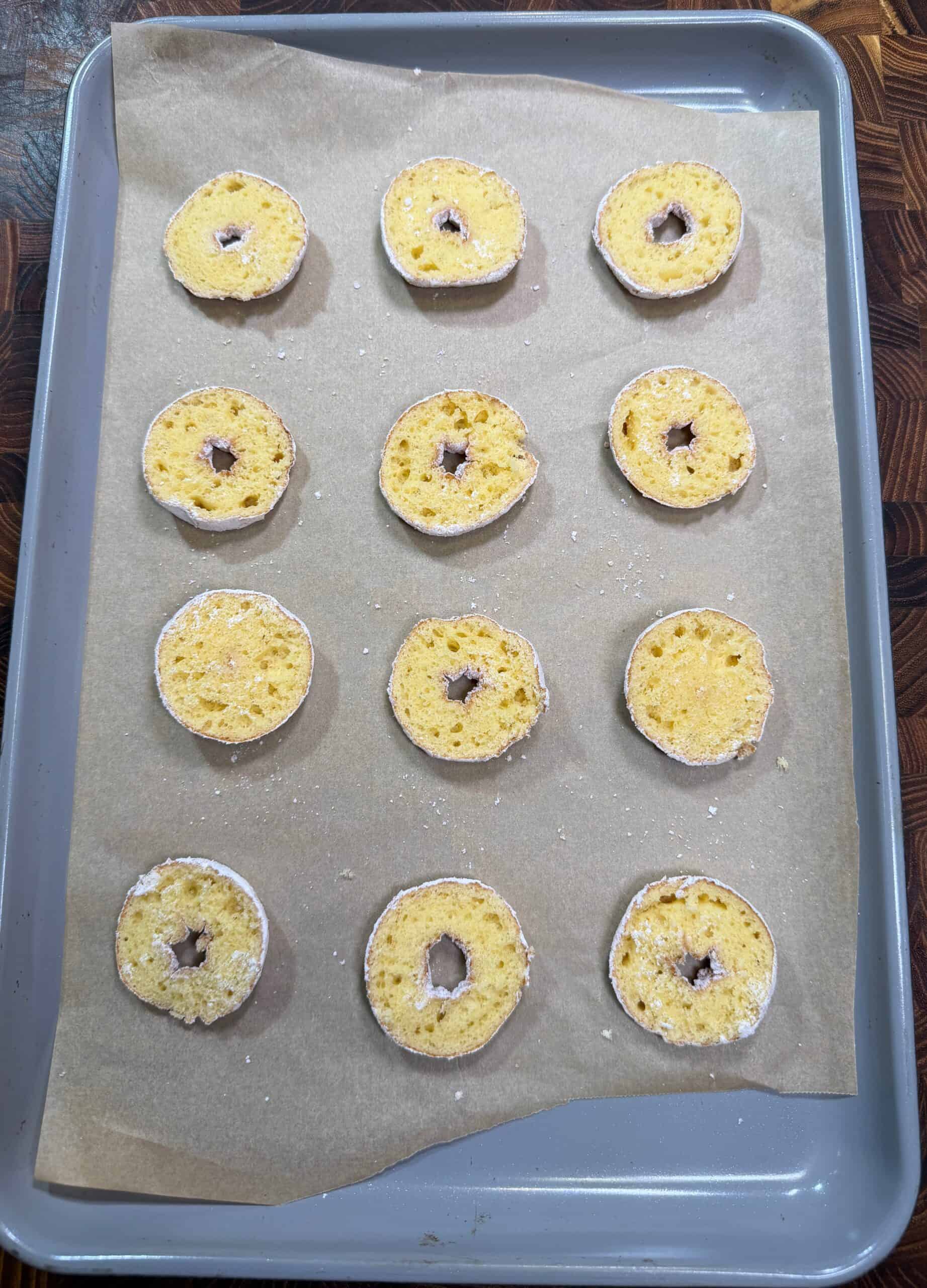 Twelve round, sliced bagel pieces with holes in the center are evenly spaced on a parchment-lined baking sheet, ready for toasting or baking. Recipe is for donut nachos