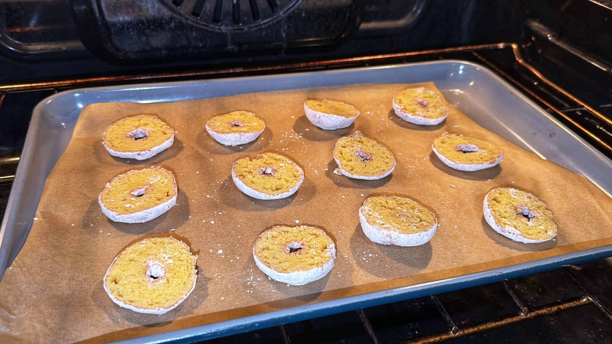 Round cookies with powdered sugar edges and a small center hole are baking on a parchment-lined baking sheet inside an oven. The cookies are evenly spaced and appear to be partially baked.