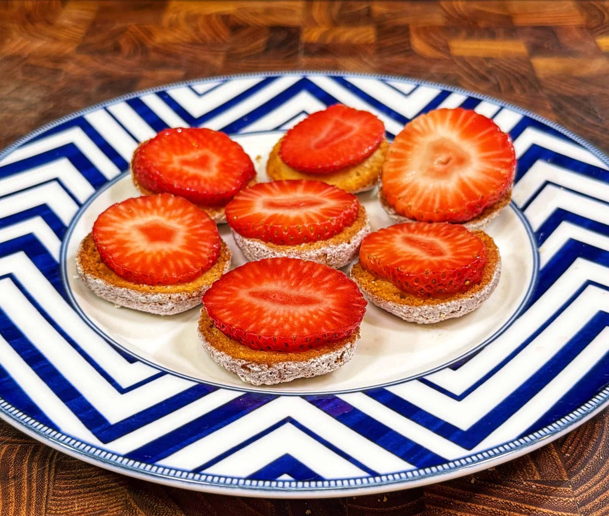 A plate with a blue-and-white zigzag pattern holds seven small round cakes, each topped with a fresh strawberry slice. The plate is set on a wooden surface.