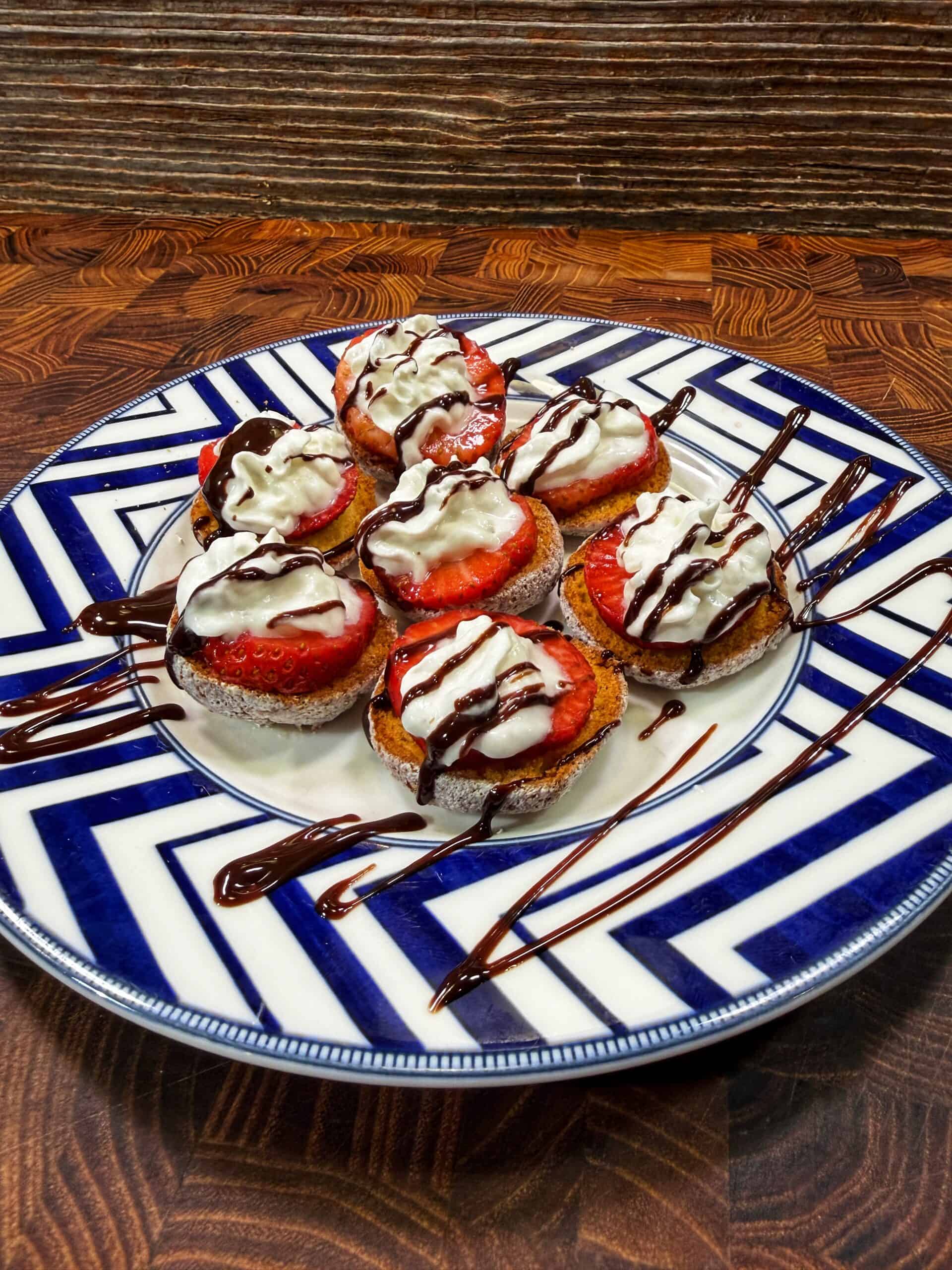 A plate of small desserts topped with sliced strawberries, whipped cream, and drizzled with chocolate sauce, arranged on a blue-and-white patterned dish placed on a wooden table.