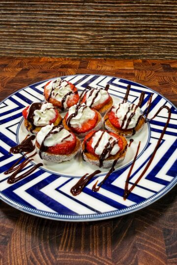 A plate of round dessert bites topped with whipped cream, sliced strawberries, and drizzled with chocolate sauce, arranged neatly on a geometric blue and white plate on a wooden surface.