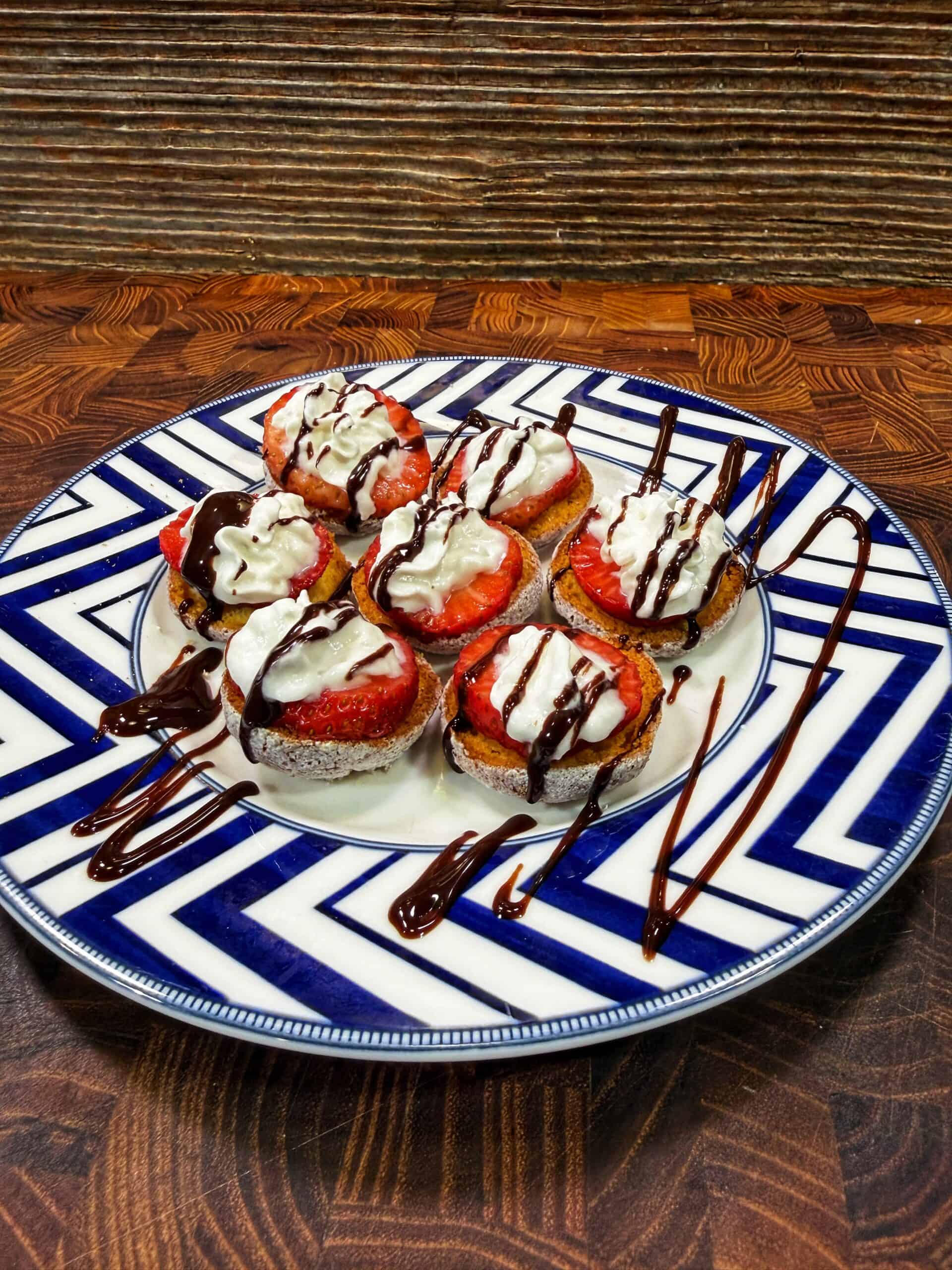 A plate of round dessert bites topped with whipped cream, sliced strawberries, and drizzled with chocolate sauce, arranged neatly on a geometric blue and white plate on a wooden surface.