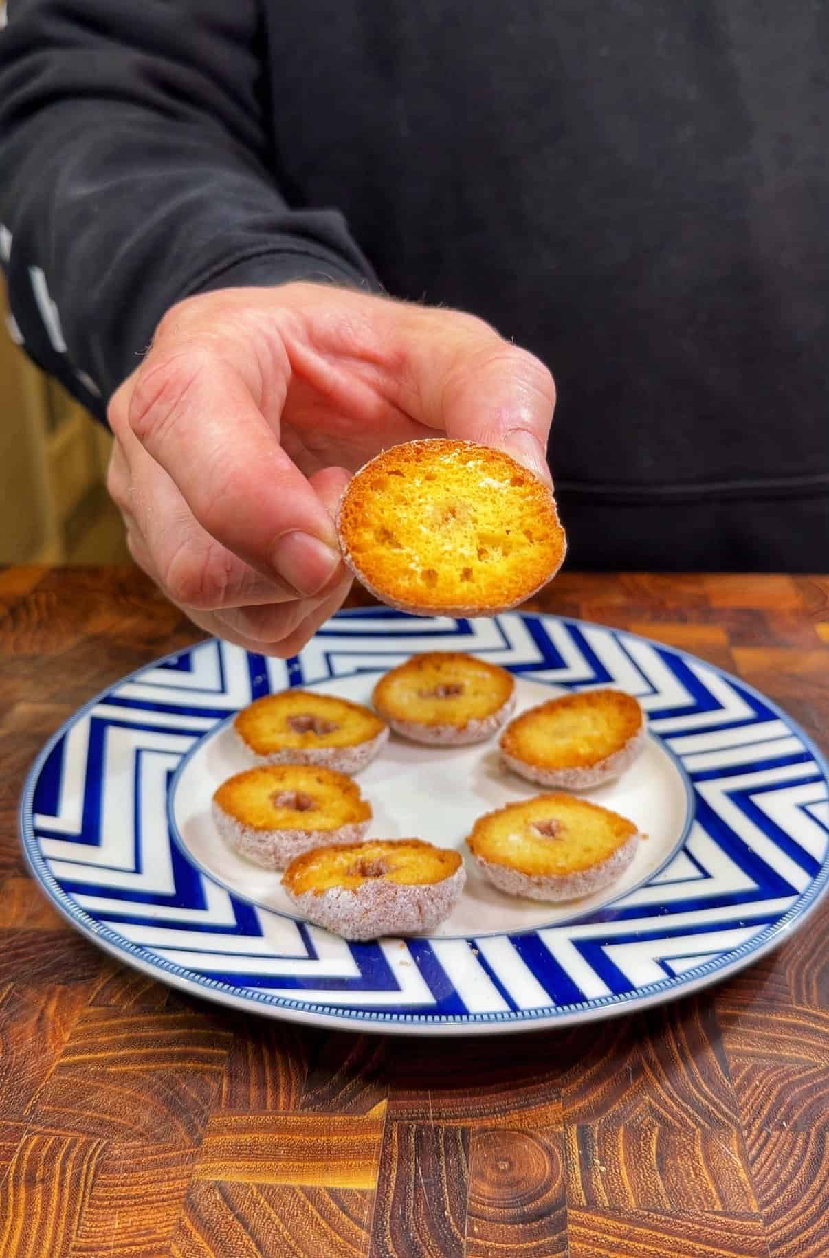 A person holds up a small, round, golden-brown baked treat over a plate with several similar treats on a wooden surface. The plate features a blue and white zigzag pattern.