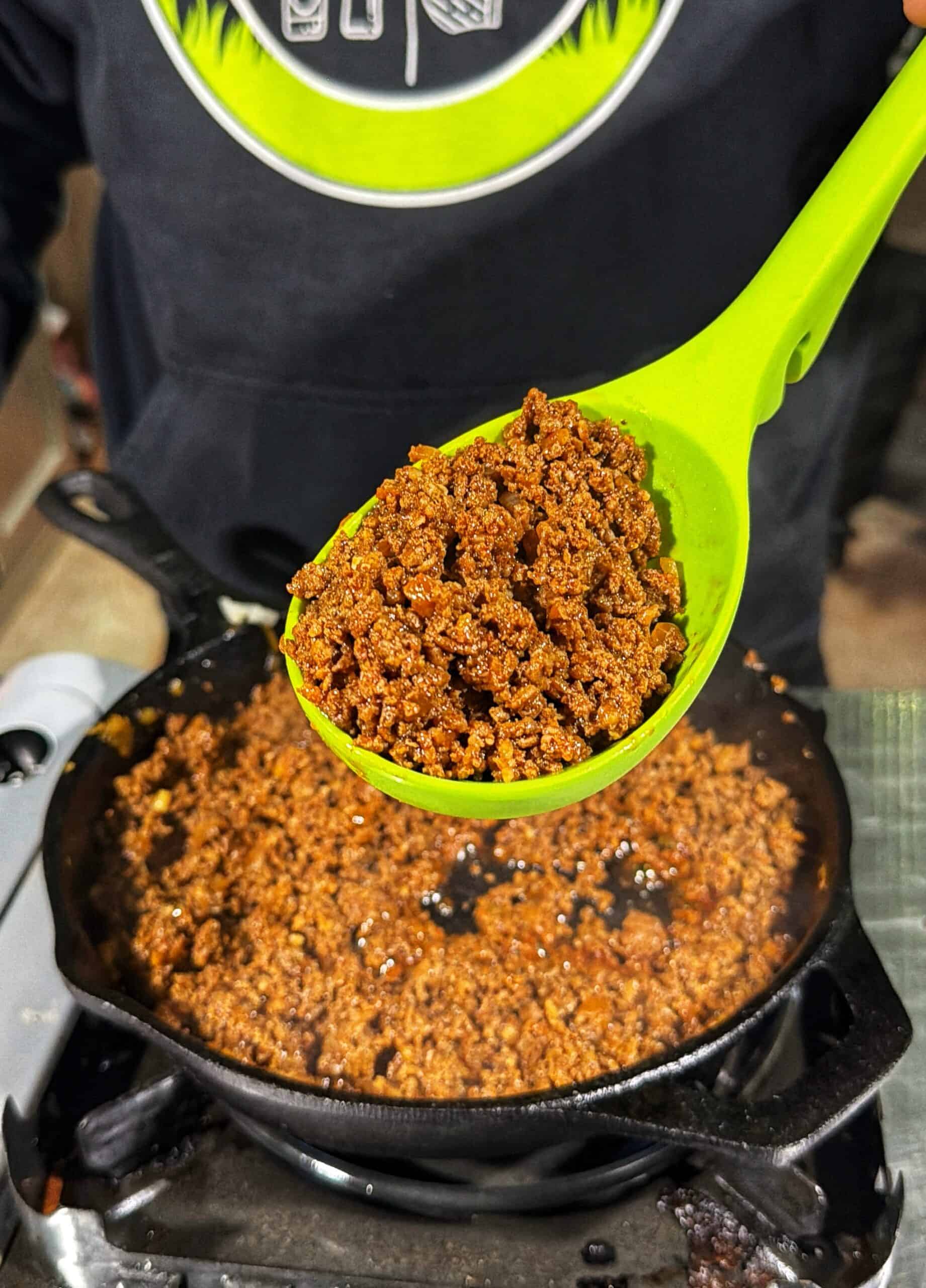 A person holds a bright green spoon filled with seasoned ground meat over a cast iron skillet containing more of the cooked meat. The person is wearing a dark hoodie.
