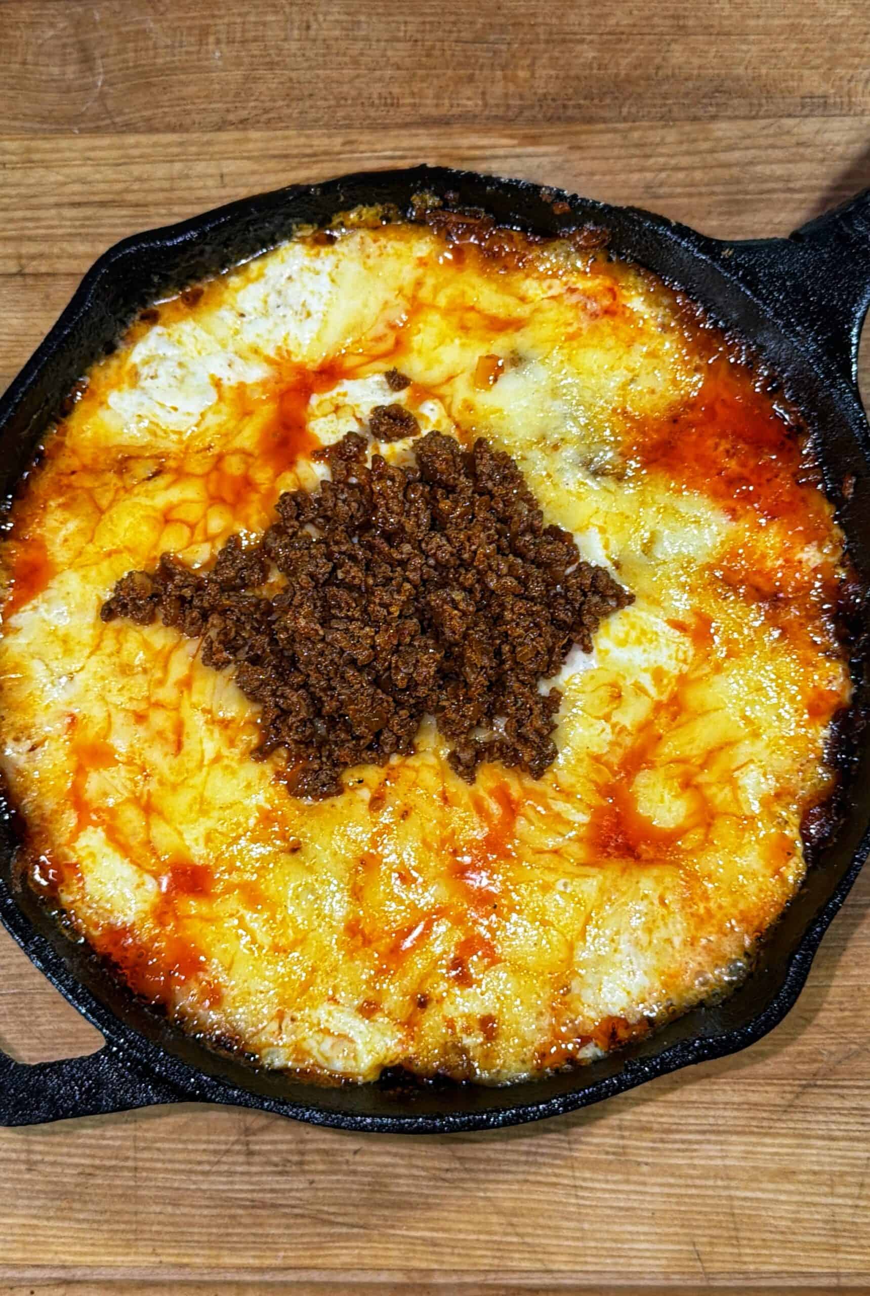 A cast iron skillet filled with melted cheese topped with cooked, crumbled chorizo, with orange oil pooling on the surface, sits on a wooden surface.