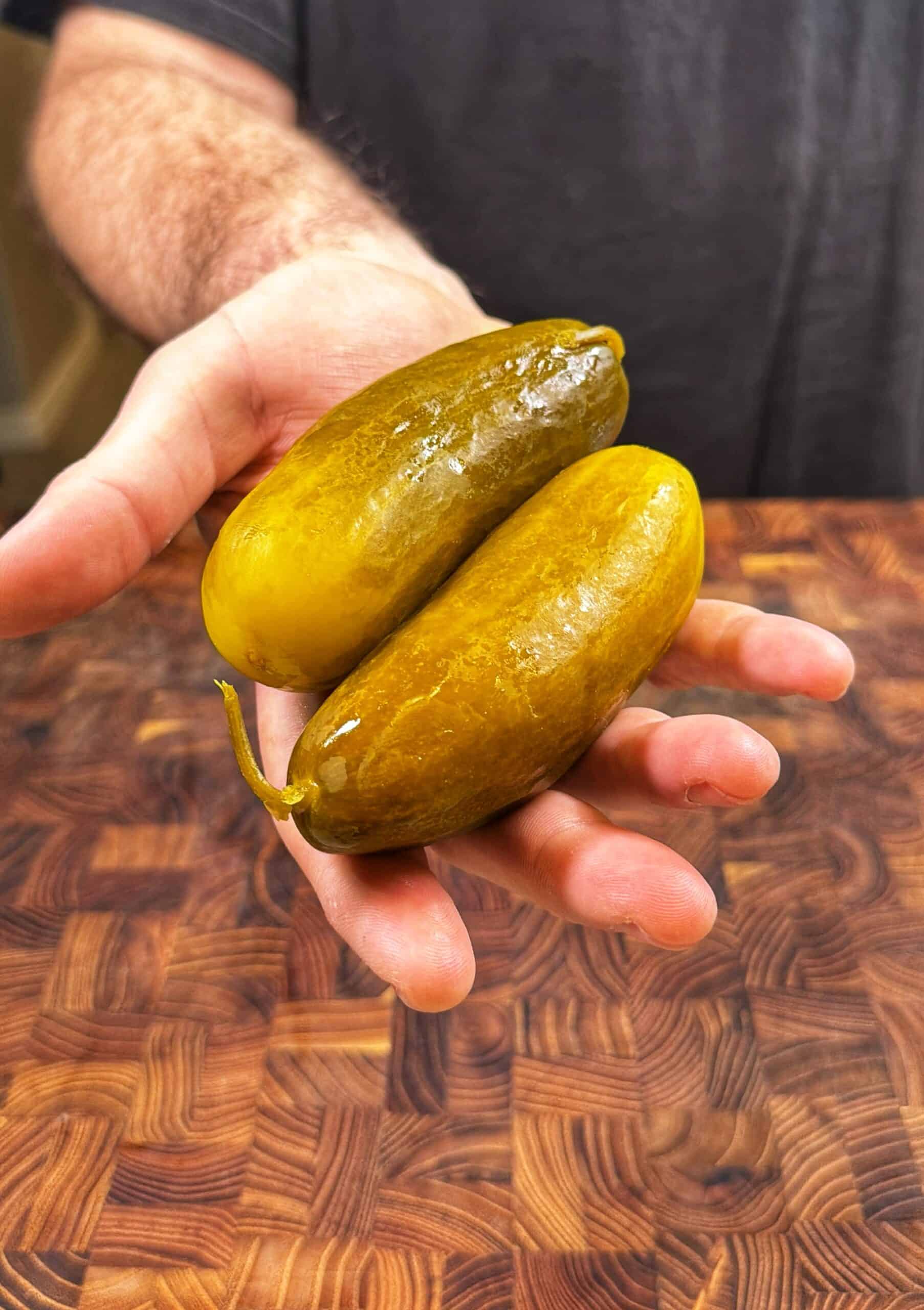 A persons hand holding two large pickles over a wooden surface.