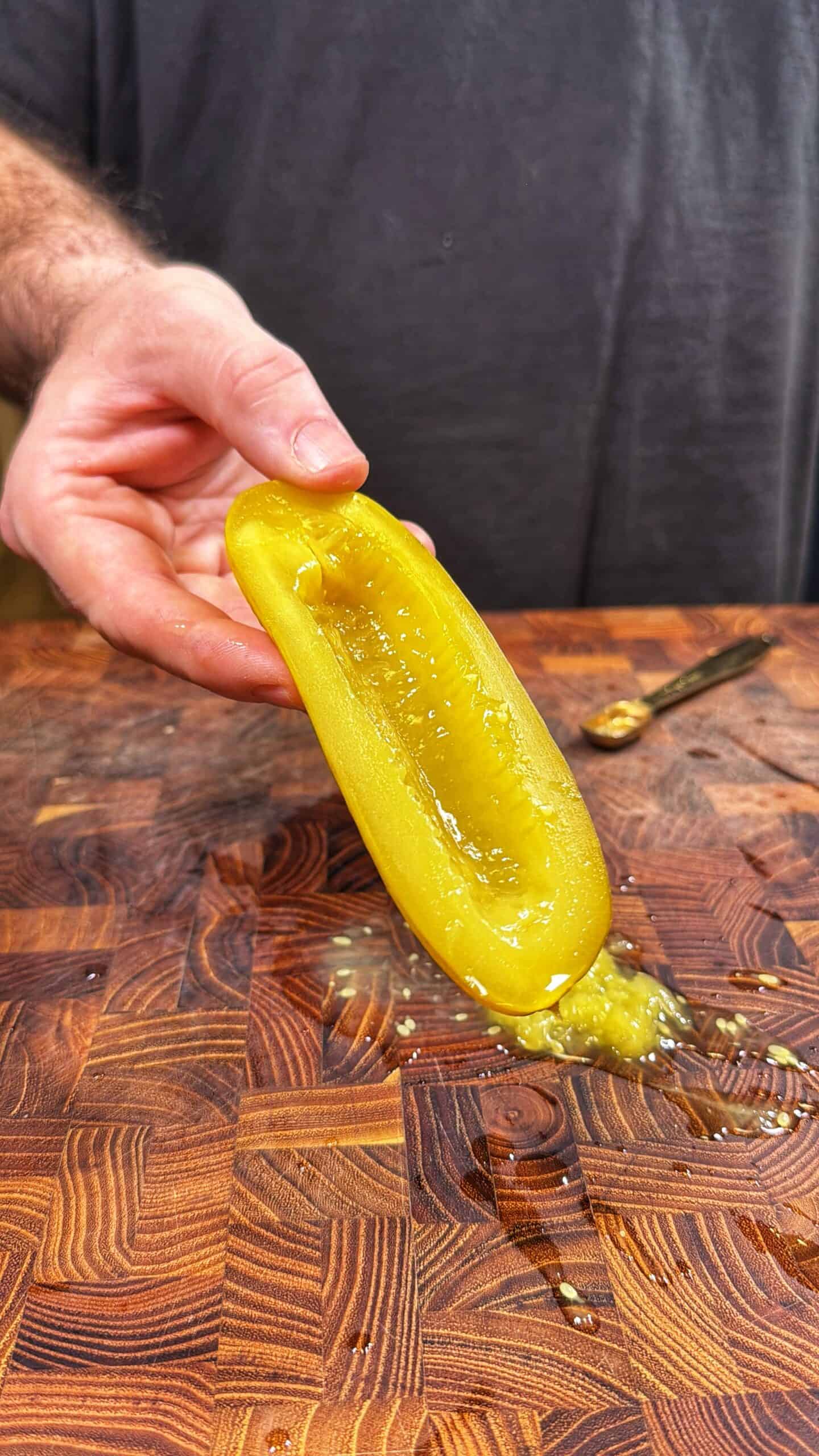 A person holds a hollowed-out pickle half over a wooden cutting board, with seeds and liquid spilling onto the surface. A small utensil lies in the background.