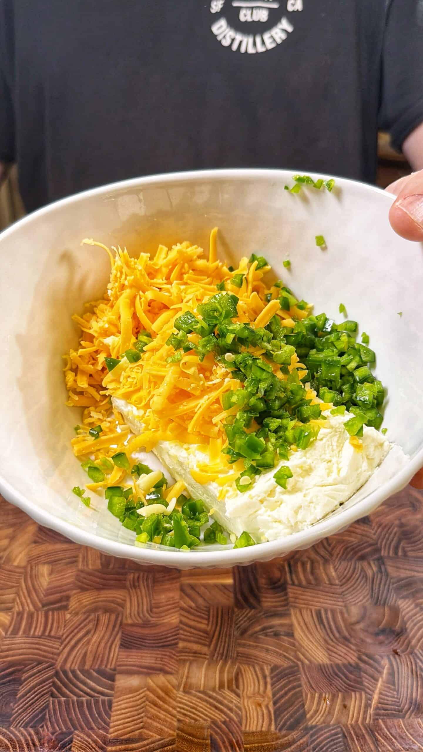 A white bowl containing shredded cheddar cheese, chopped green onions, and a block of cream cheese is held in front of a person wearing a black shirt. The bowl rests on a wooden surface.