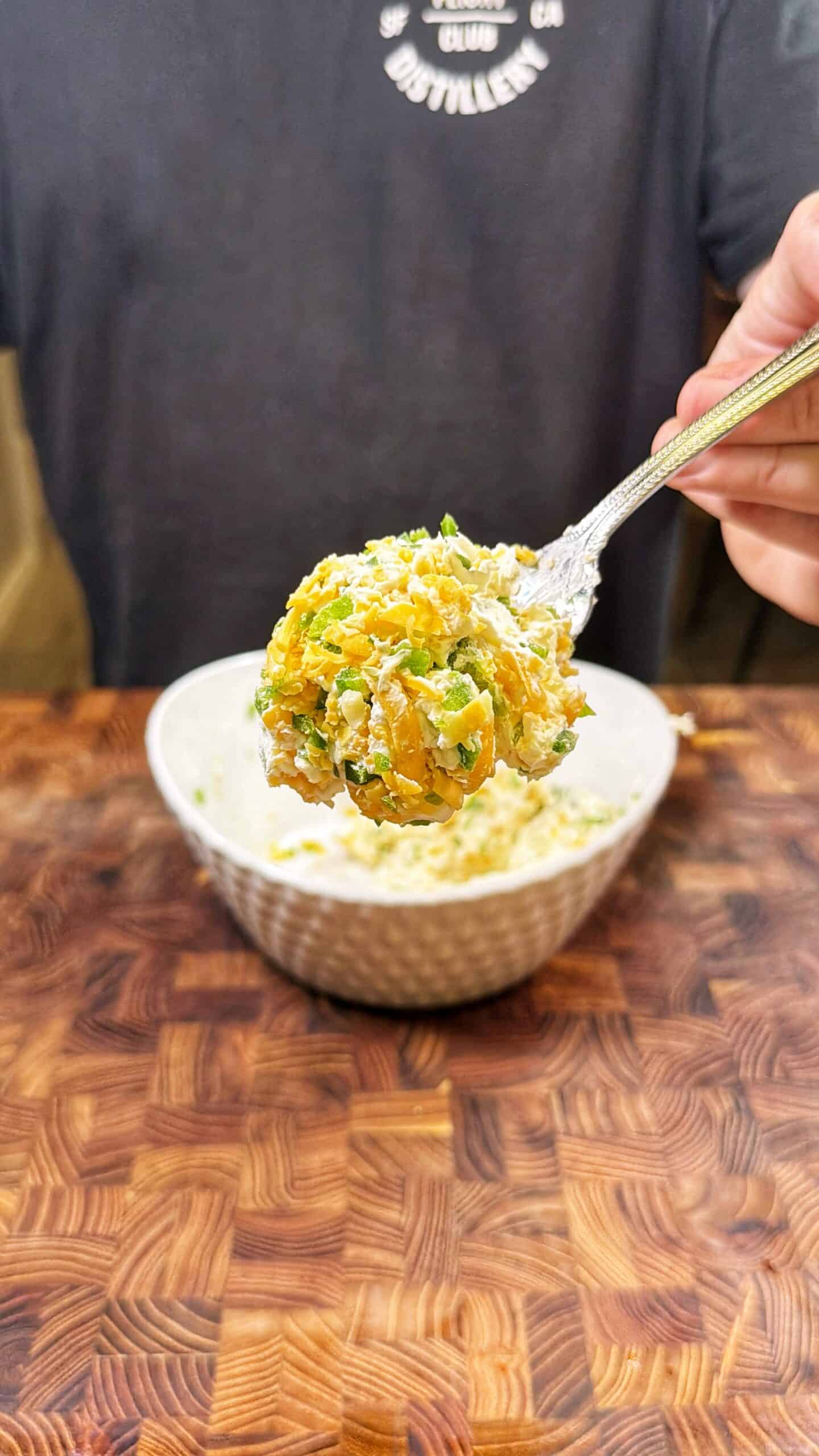 A hand holds a spoonful of mixed salad with corn, green herbs, and white dressing above a white bowl on a wooden table.