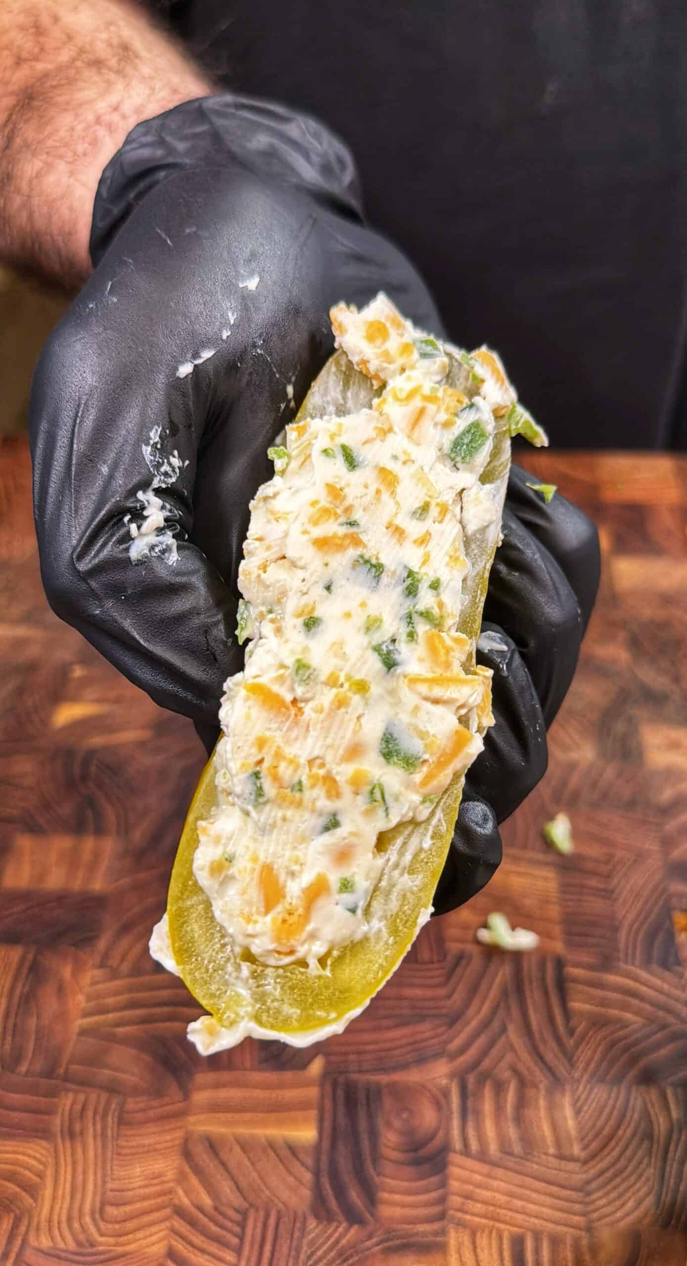 A hand wearing a black glove holds a sliced pickle filled with a creamy mixture containing chopped herbs and orange bits, over a wooden cutting board. Recipe is for a smoked pickle poppers.