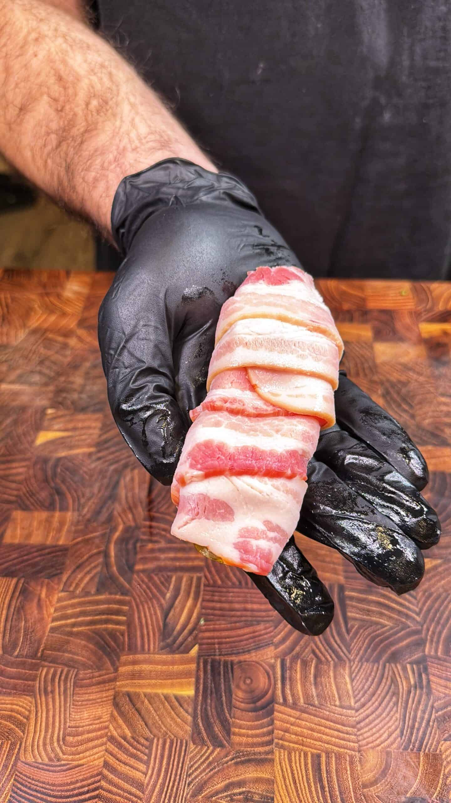 A person wearing a black glove holds a piece of raw meat wrapped in bacon over a patterned wooden surface.