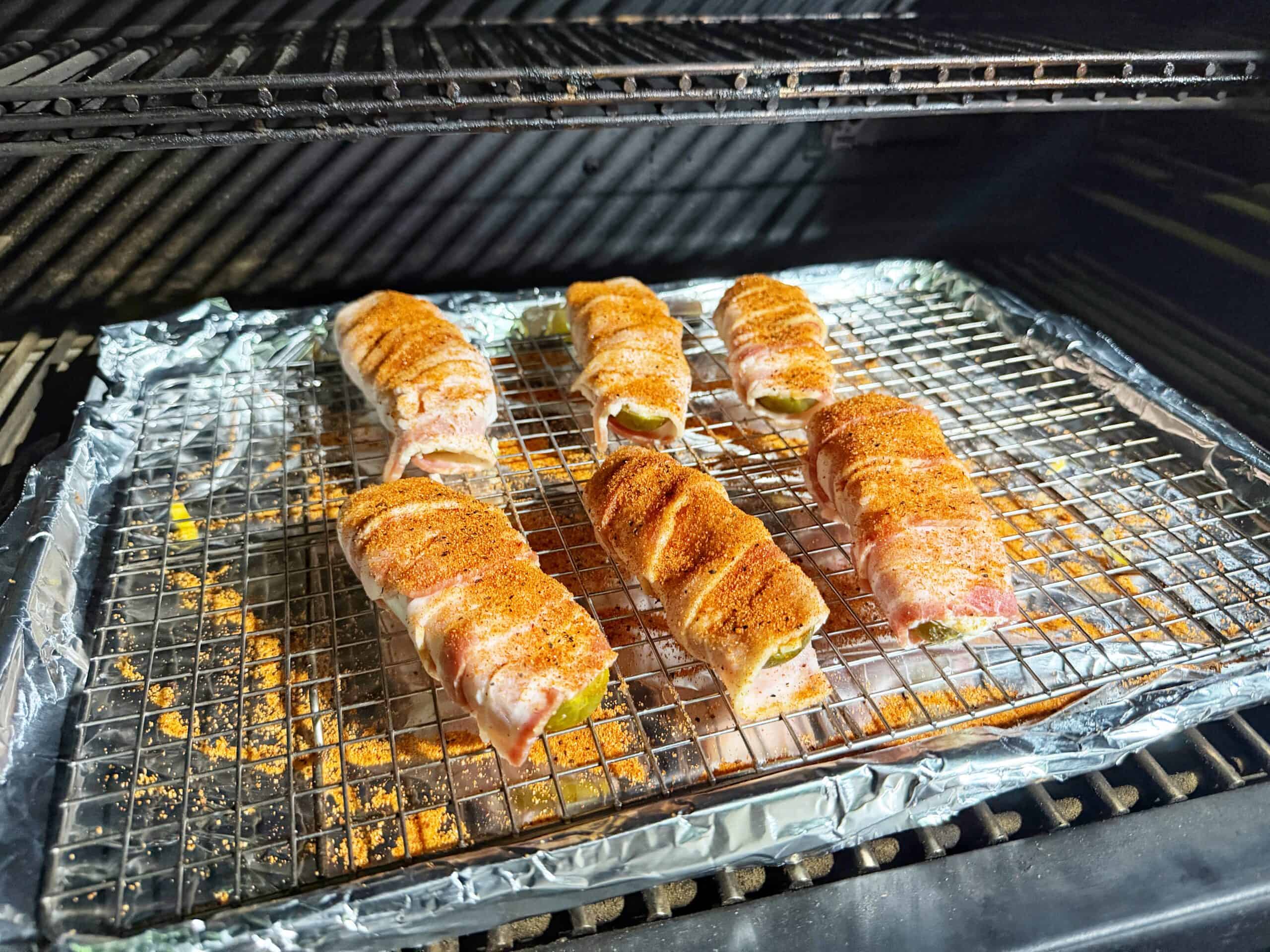 Six bacon-wrapped, seasoned jalapeño poppers are arranged on a foil-lined rack inside an oven or grill, ready to be cooked.