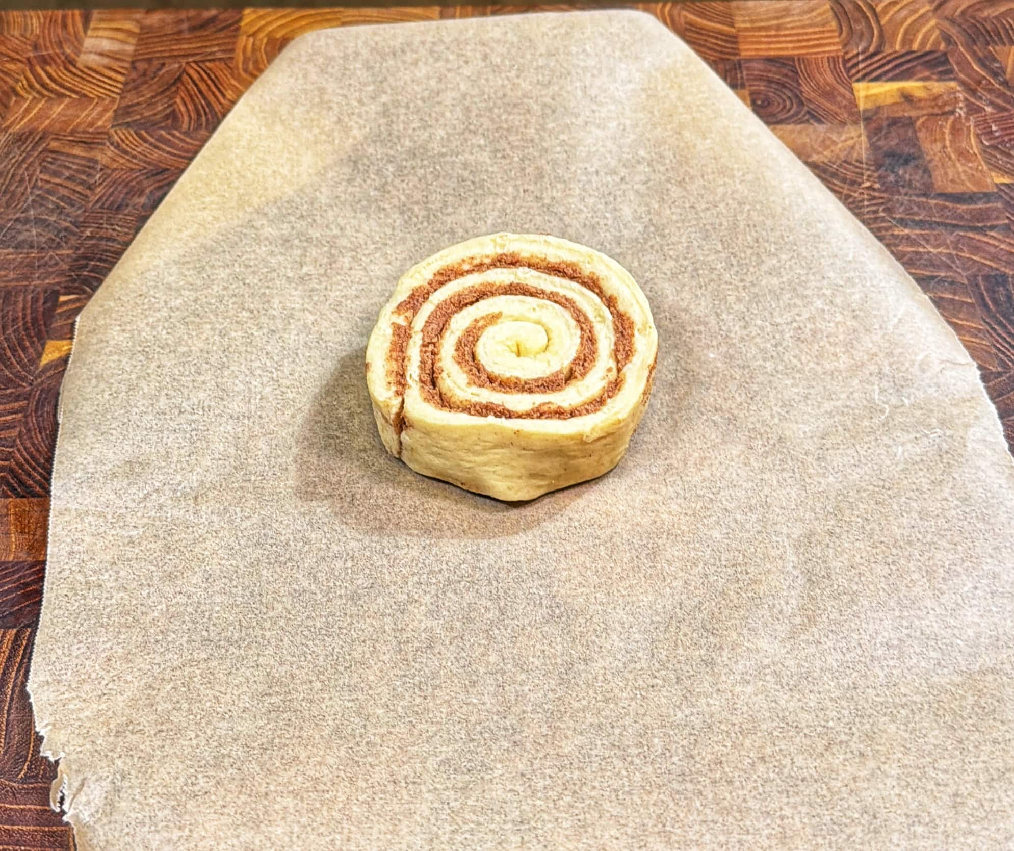 A single unbaked cinnamon roll sits on a sheet of parchment paper atop a wooden surface, showing its spiral layers of dough and cinnamon filling.