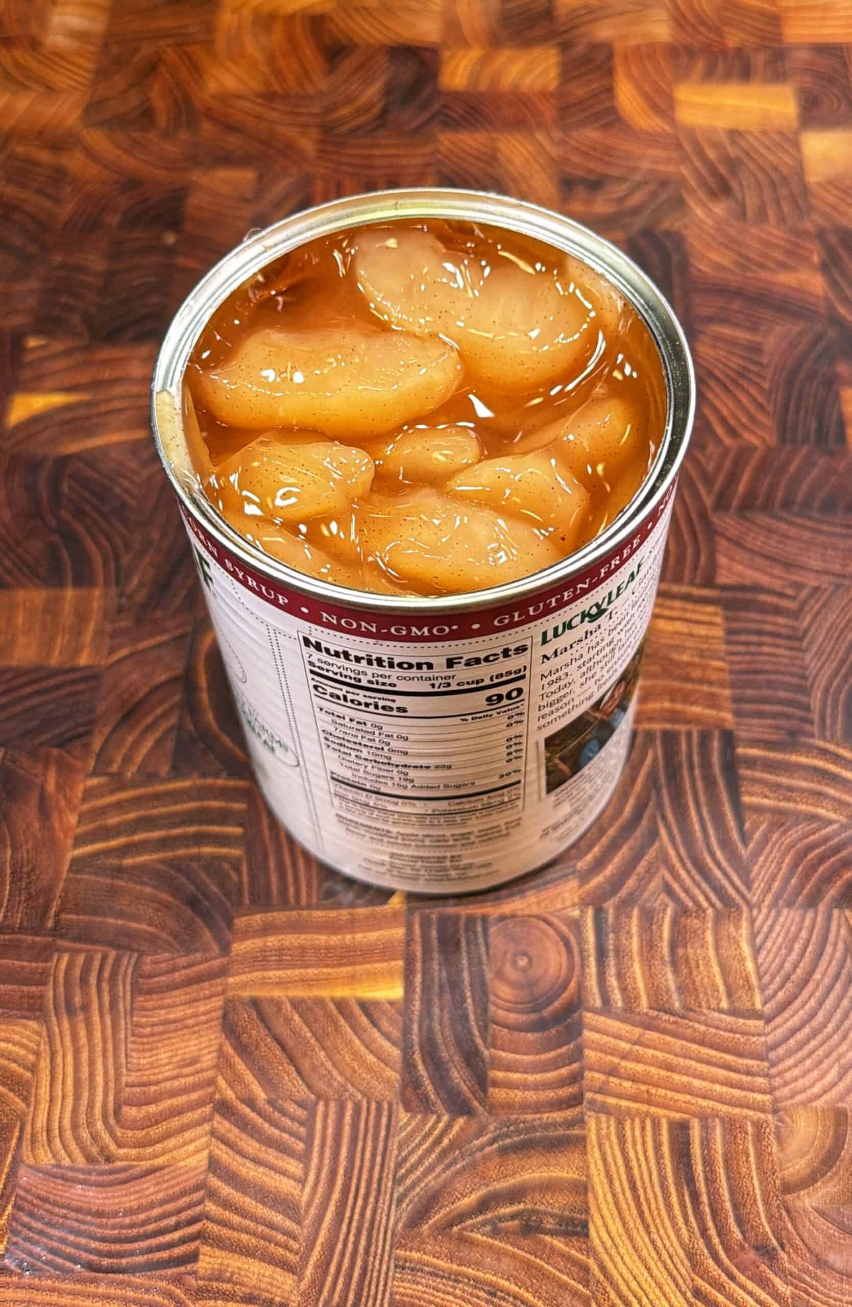 An open can of sliced peaches in syrup sits on a wooden, patterned countertop. The can’s lid is completely removed, and the peach slices are visible above the syrup. Nutritional information is partially visible on the label.