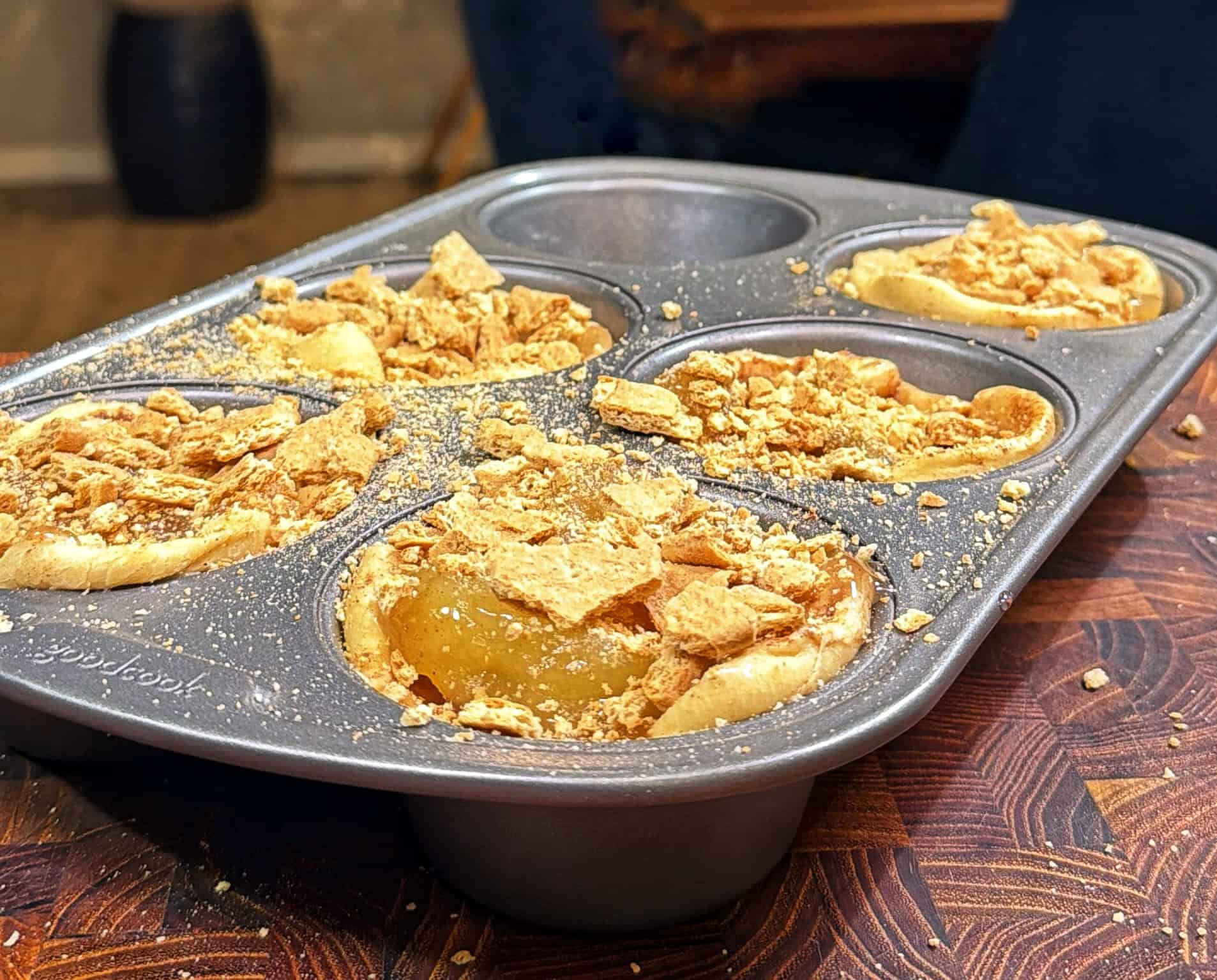 A muffin tin filled with partially baked dessert cups topped with crumbled graham crackers, sitting on a wooden surface. Some cups have filling visible under the crumbs.