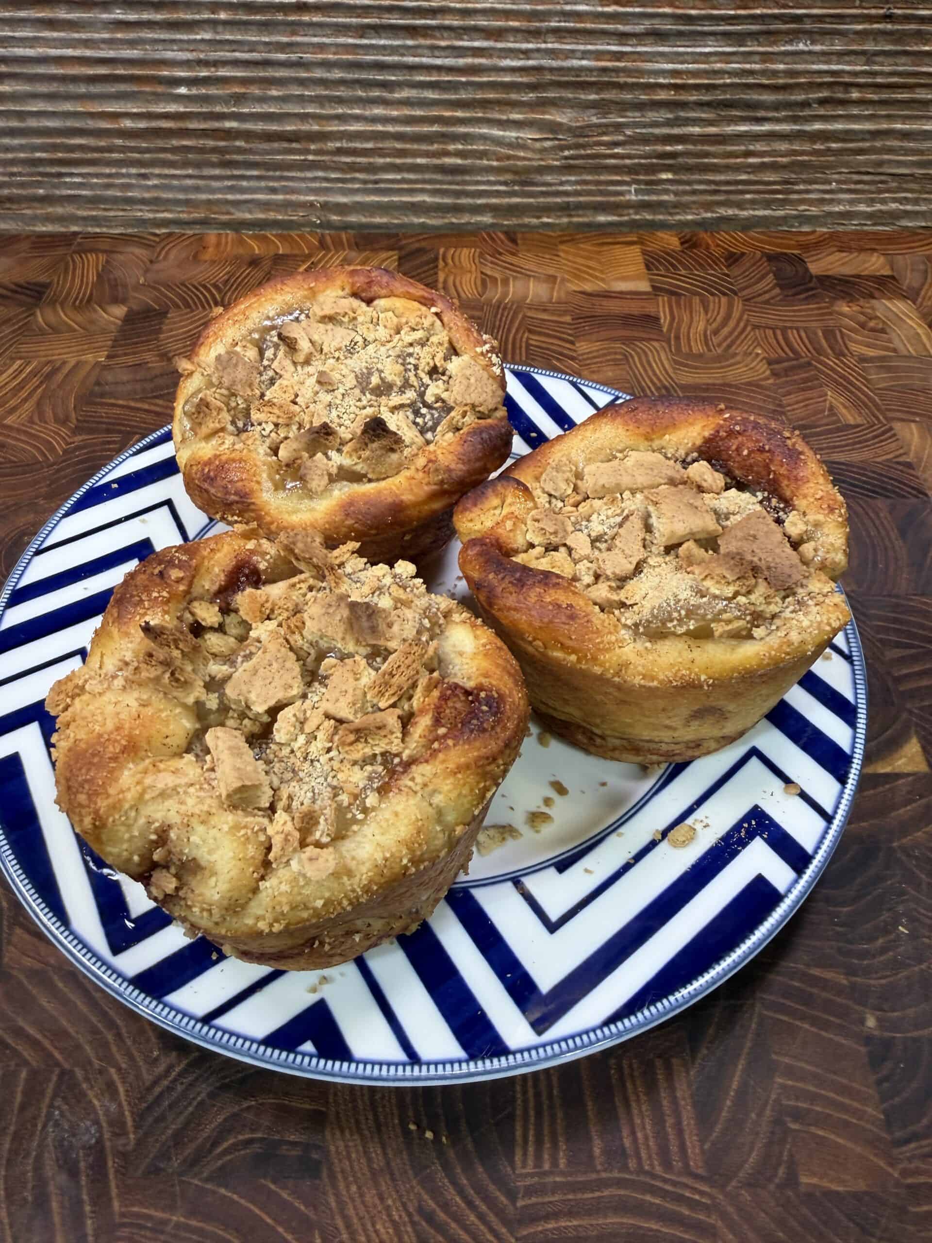 Three golden-brown muffins topped with crushed graham crackers sit on a blue and white geometric-patterned plate on a wooden surface with a textured backdrop.