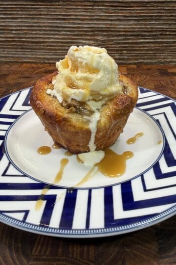 A warm pastry topped with a scoop of vanilla ice cream and drizzled with caramel sauce sits on a patterned blue and white plate on a wooden surface.