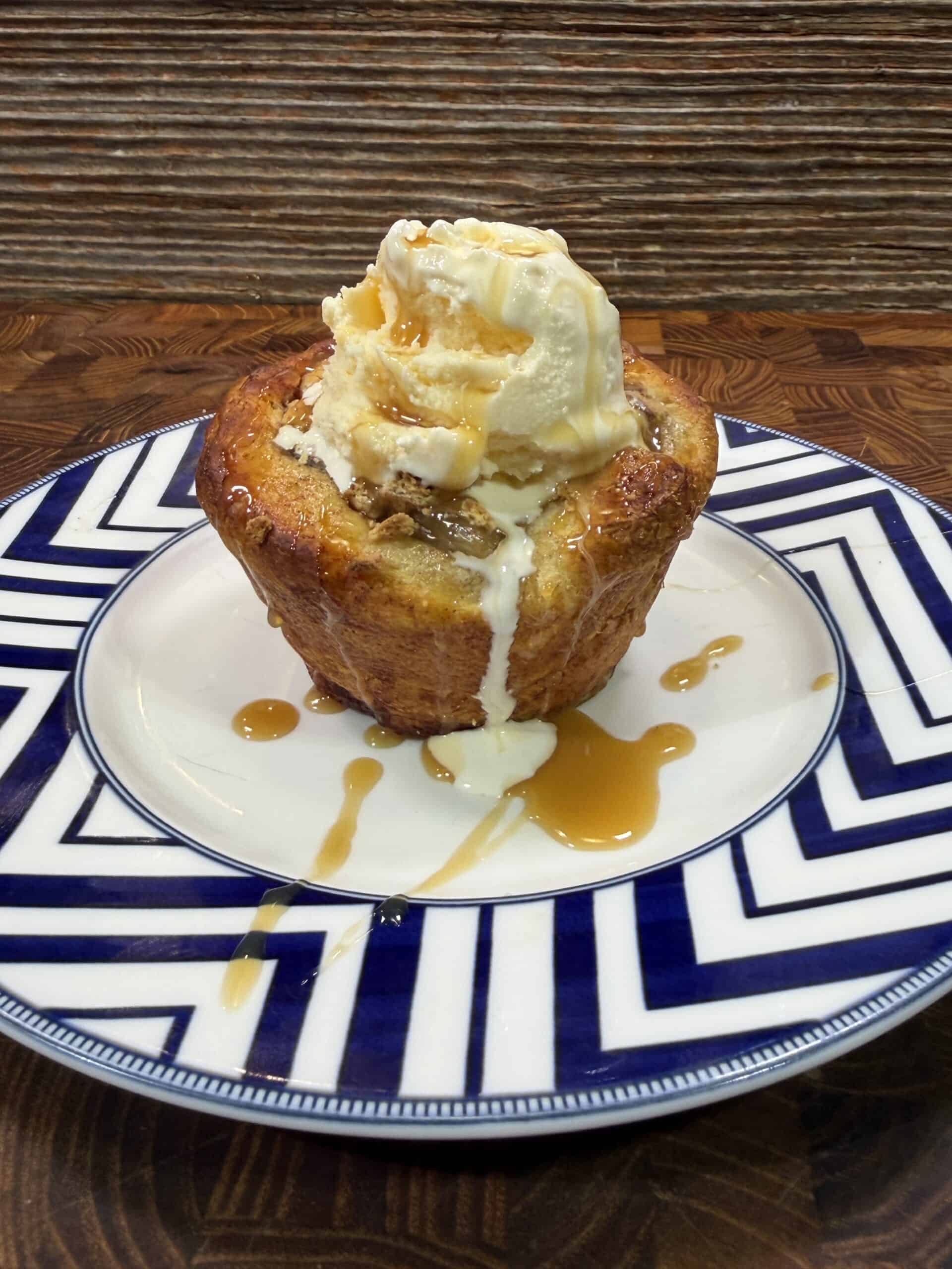 A warm apple pie cinnamon roll muffin topped with a scoop of vanilla ice cream and drizzled with caramel sauce sits on a patterned blue and white plate on a wooden surface.