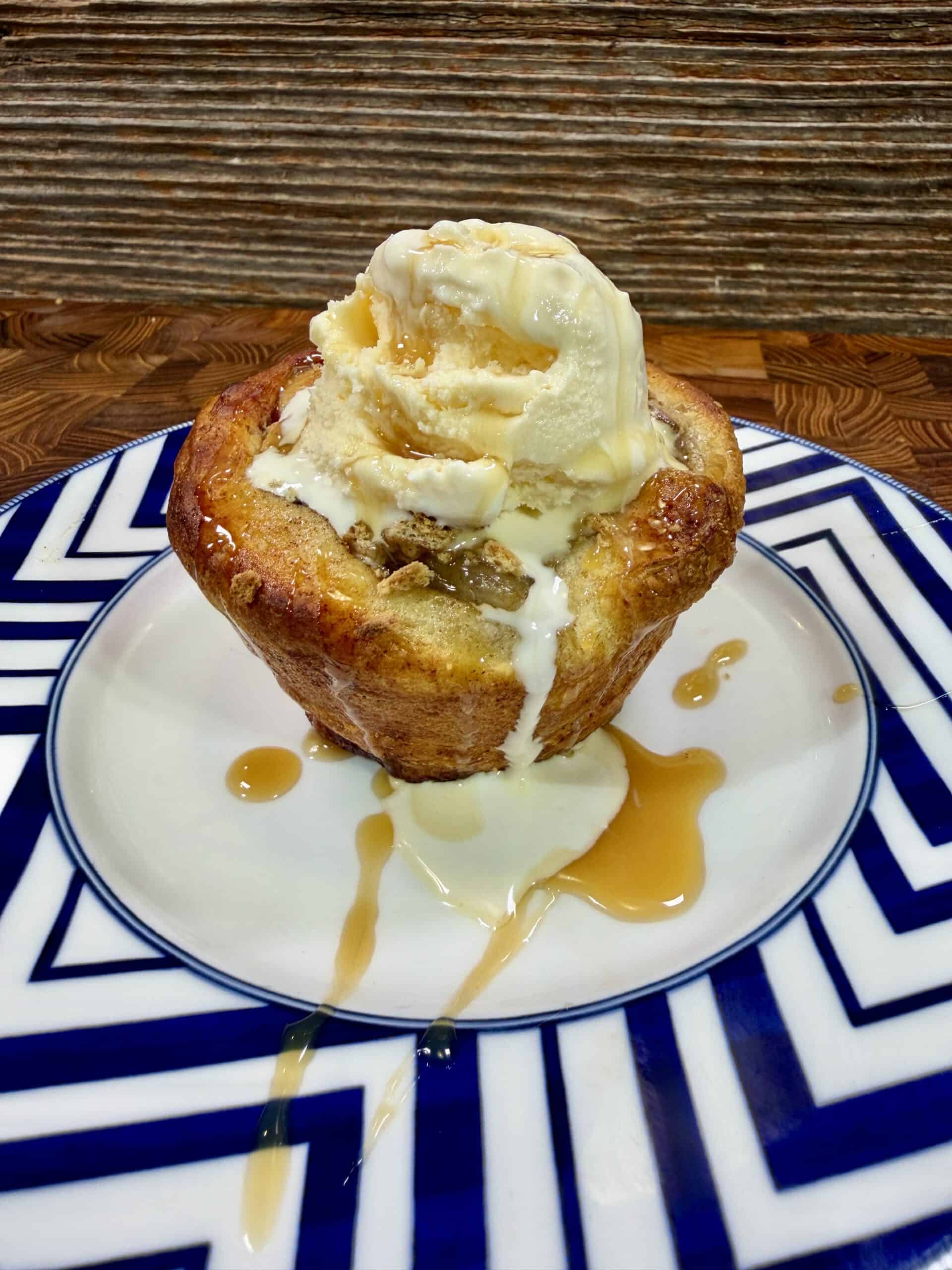 A baked dessert topped with a scoop of vanilla ice cream and caramel sauce, served on a white plate with a blue geometric pattern. The ice cream is melting, and caramel drips onto the plate.