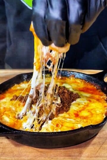 A gloved hand lifts melted, stretchy cheese from a cast iron skillet filled with ground meat and bubbling, golden cheese on a wooden surface.