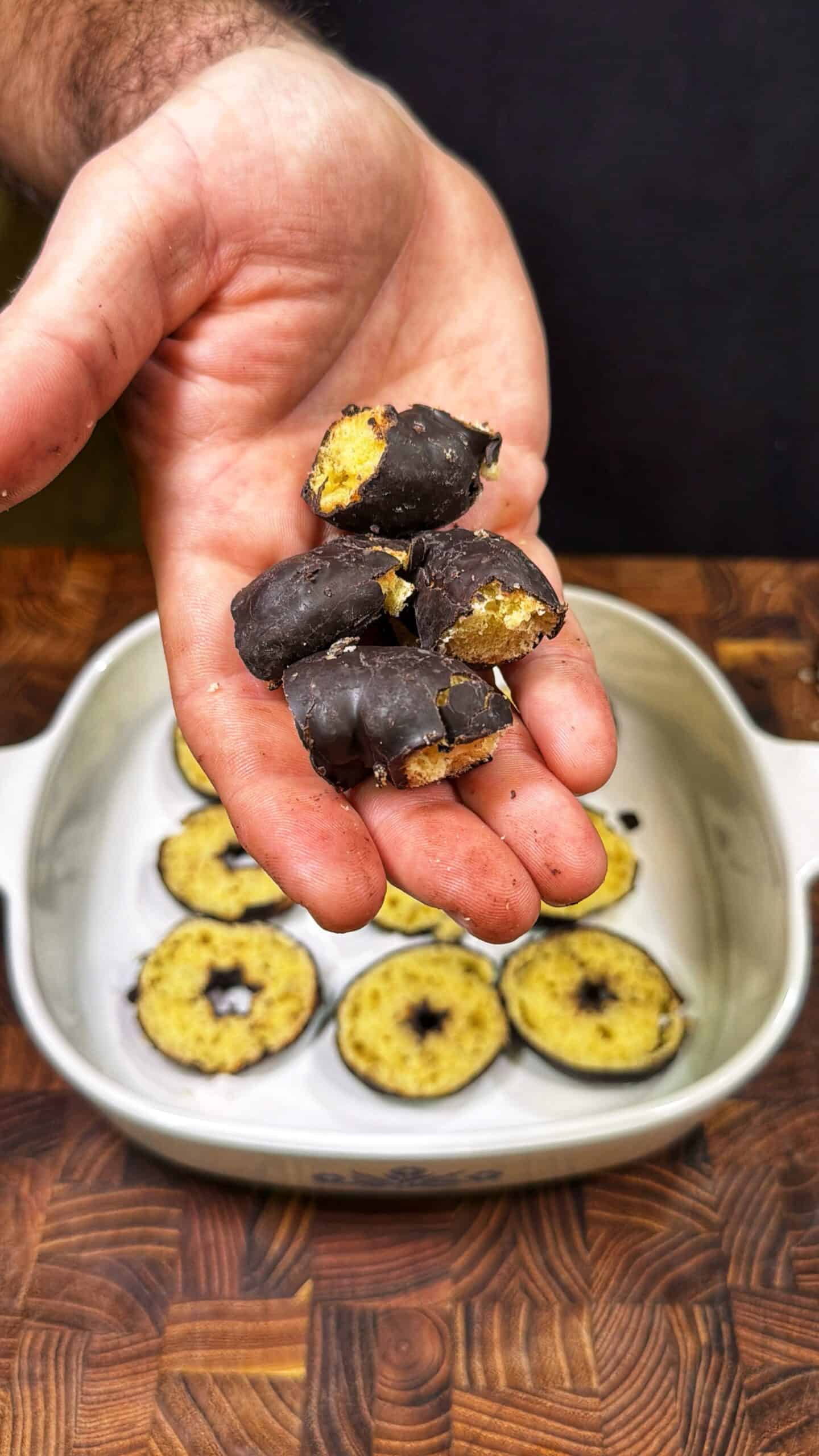 A hand holds several small, round, chocolate-coated treats with a yellow filling. In the background, more of the same treats are visible in a white dish on a wooden surface.