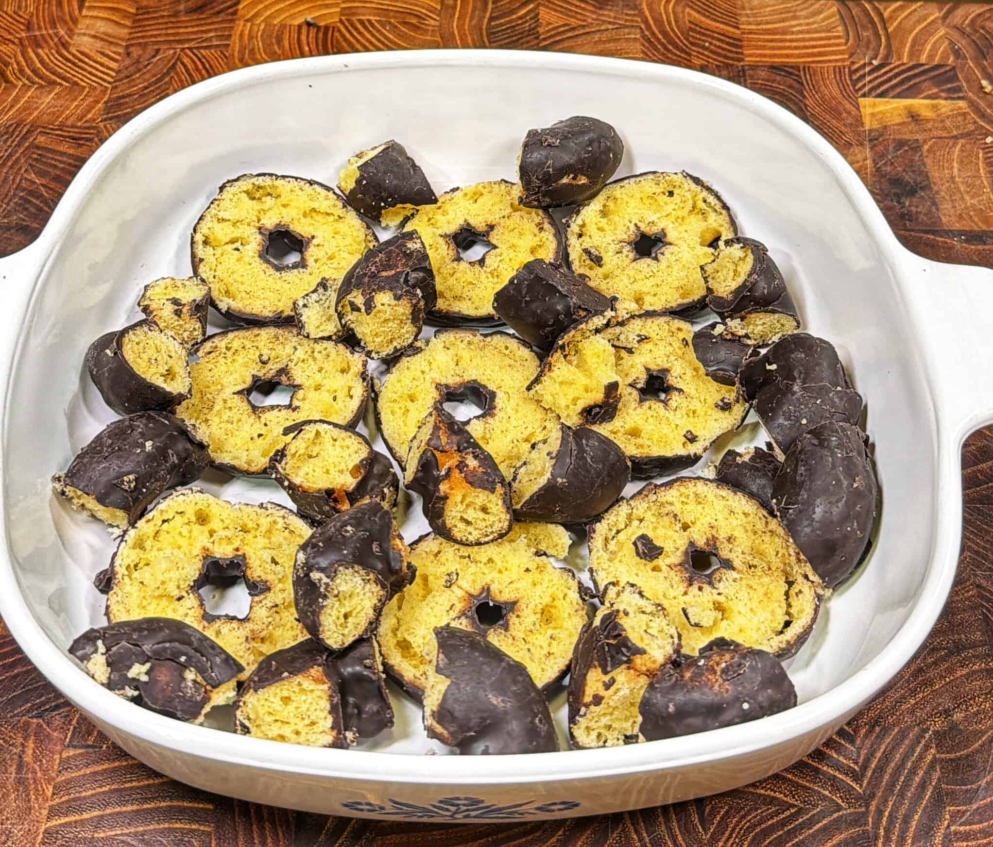 A white casserole dish holds crumbled chocolate-covered cookies, arranged in a single layer on a wooden surface. The cookies are ring-shaped with chocolate coating on the outer edge.