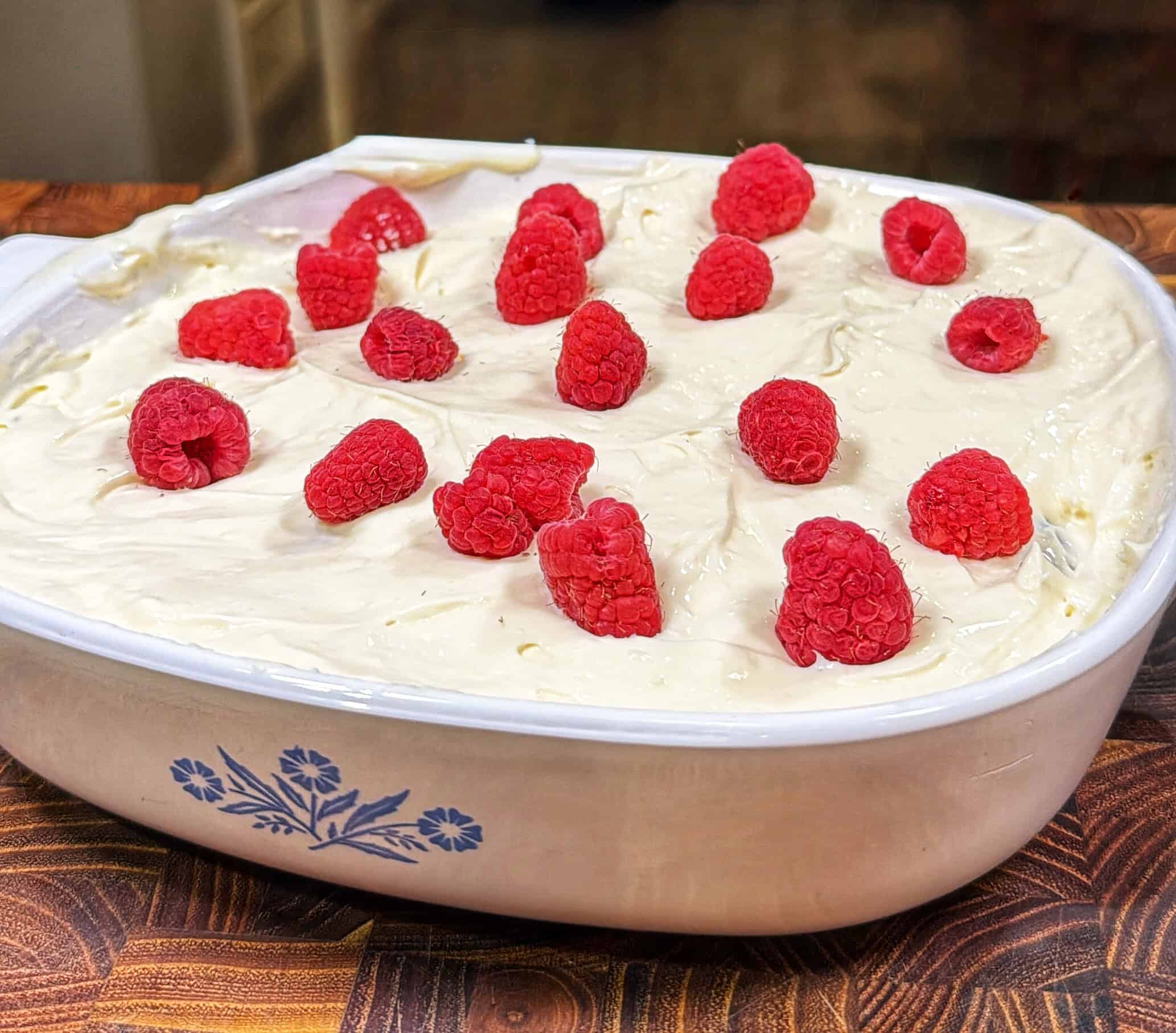 A white casserole dish filled with donut crusted cheesecake topped with fresh raspberries sits on a wooden surface. The dish has a blue floral pattern on the side.