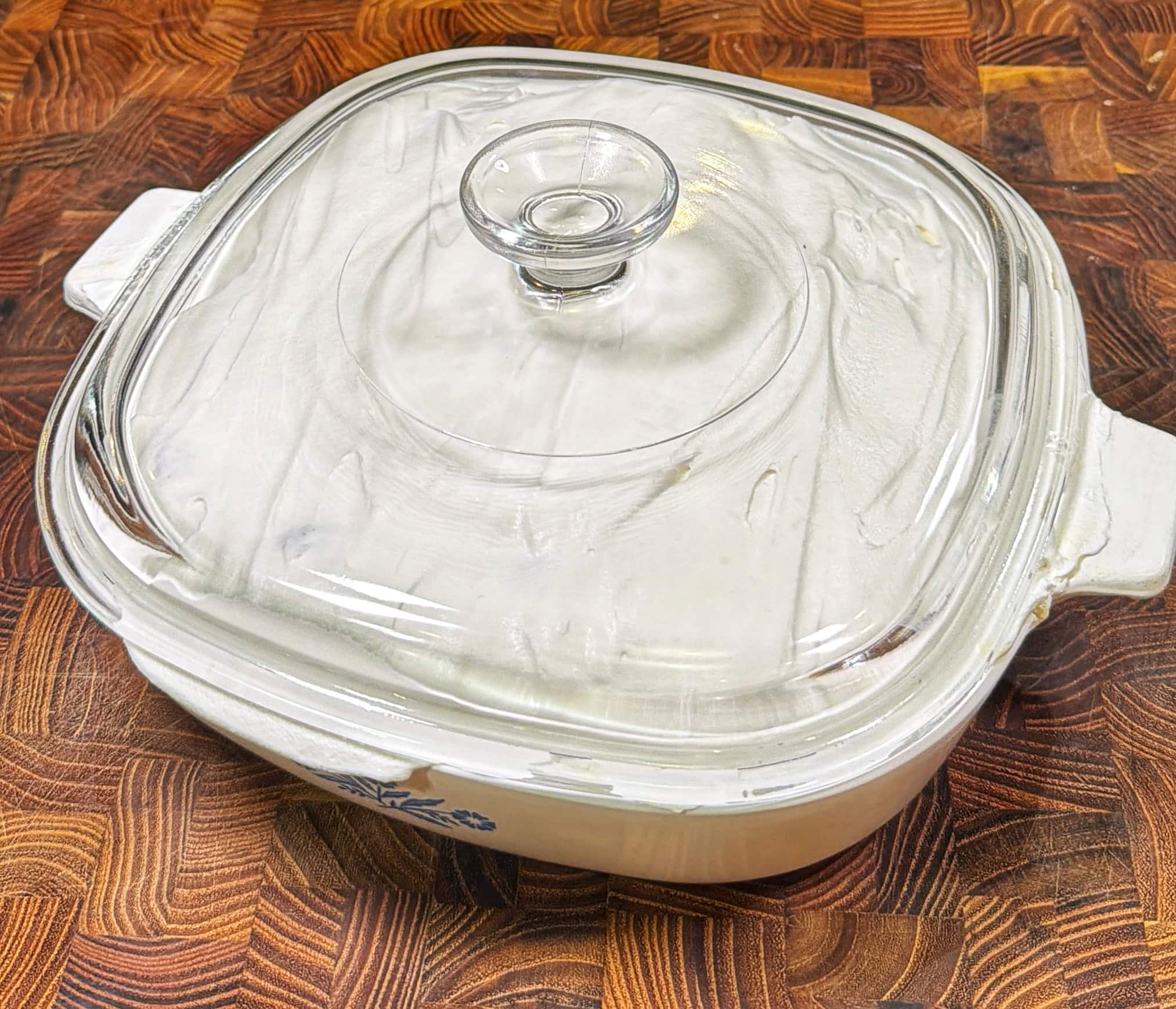 A square white casserole dish with a clear glass lid sits on a wooden countertop. The dish has handles on each side and features a blue floral design on the front.