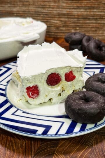 A slice of creamy dessert with visible raspberries inside, topped with whipped cream, sits on a blue and white plate next to three chocolate-covered mini donuts. The rest of the dessert is in a dish in the background.