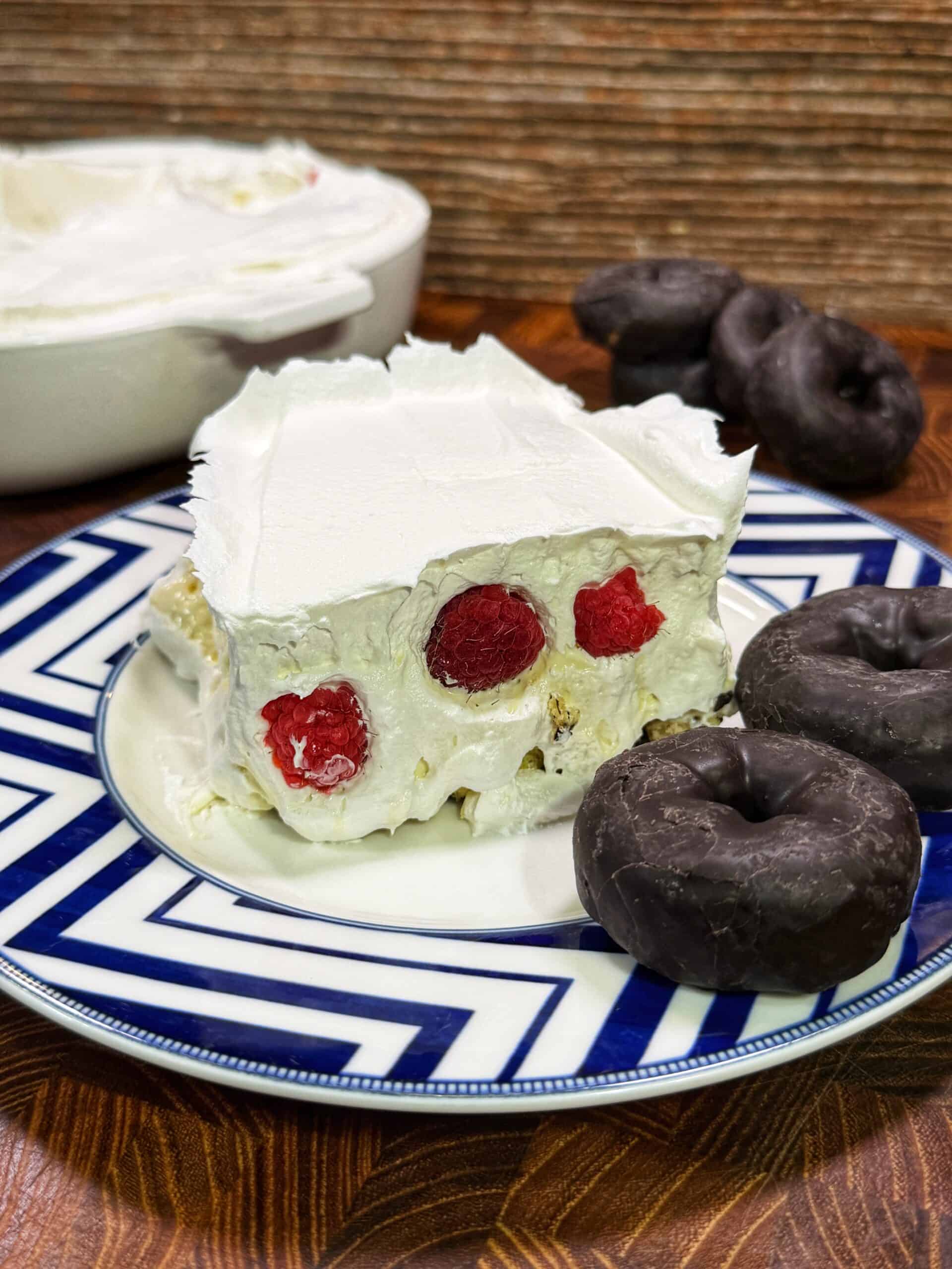A slice of creamy dessert with visible raspberries inside, topped with whipped cream, sits on a blue and white plate next to three chocolate-covered mini donuts. The rest of the dessert is in a dish in the background.