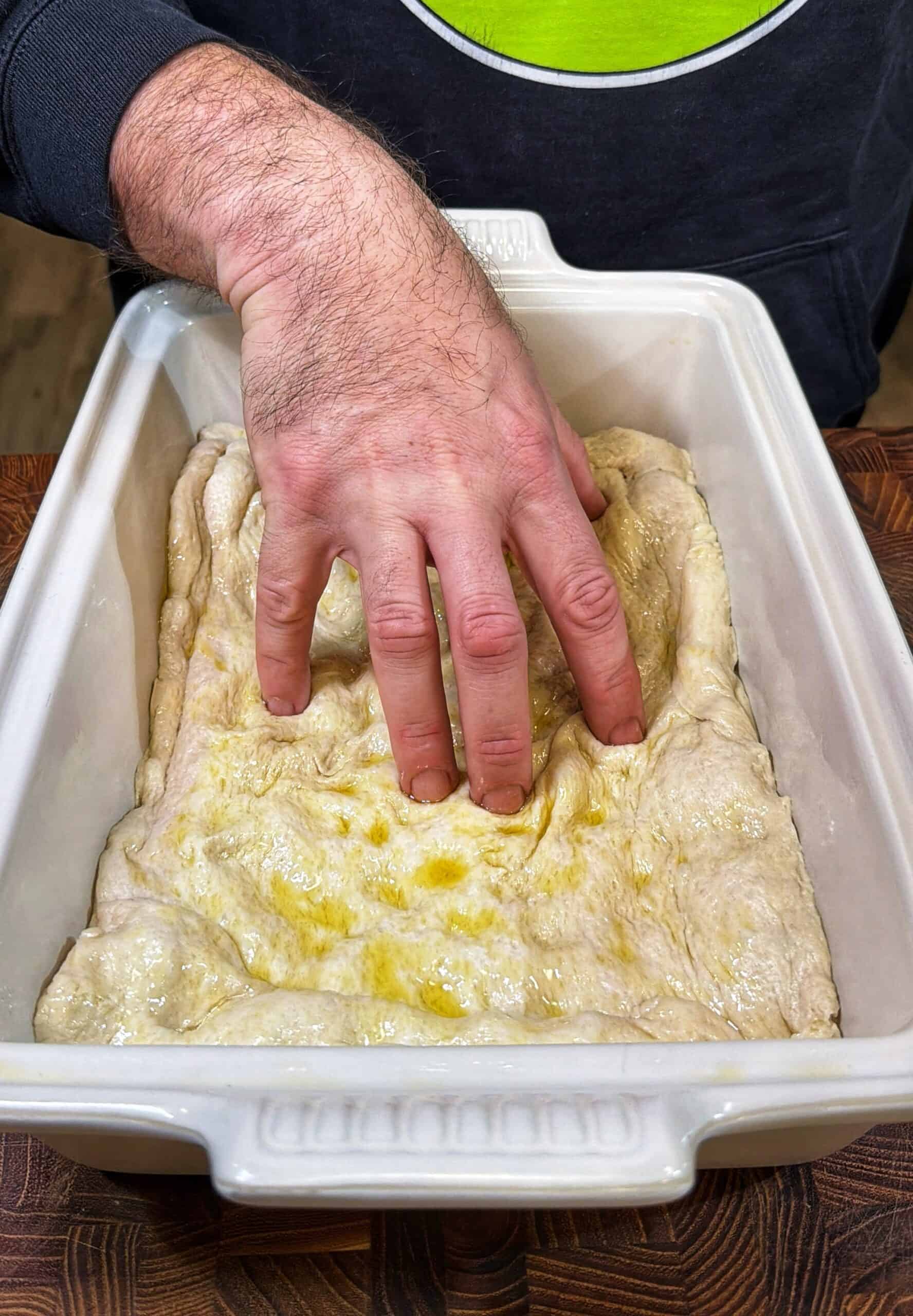 A person presses their fingers into focaccia dough in a rectangular white baking dish, preparing it for baking. The dough is drizzled with olive oil and the scene is set on a wooden surface.