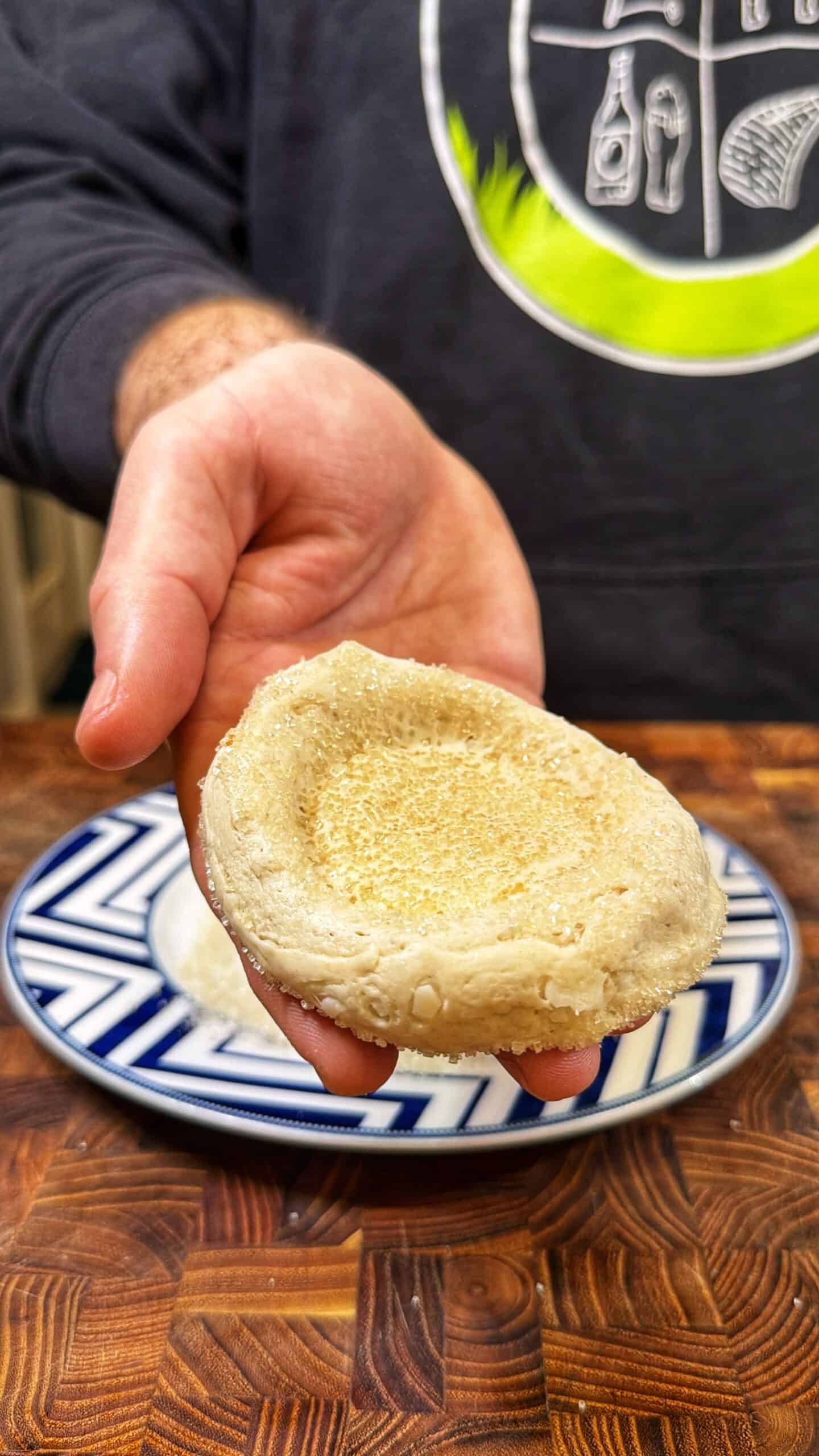 A hand holds a raw, round dough with an indented center over a patterned plate on a wooden surface. The person is wearing a dark sweatshirt with a green and white design.