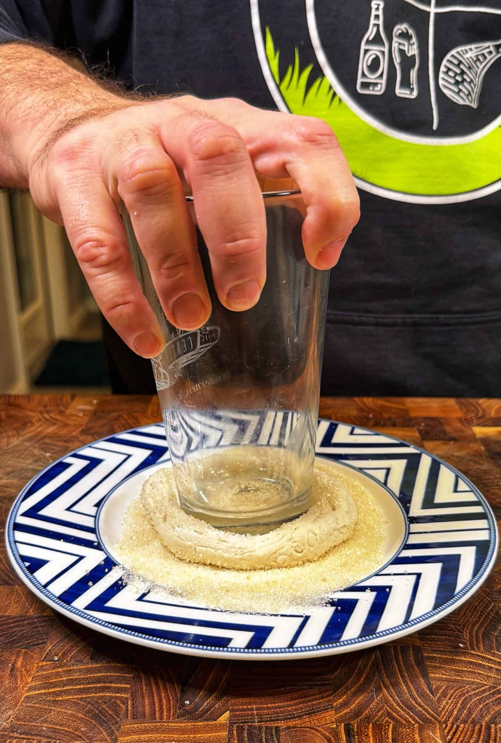 A hand presses a glass upside-down onto a round piece of dough on a plate covered with sugar, on a wooden surface. The person is wearing a dark shirt with a green and white graphic design.