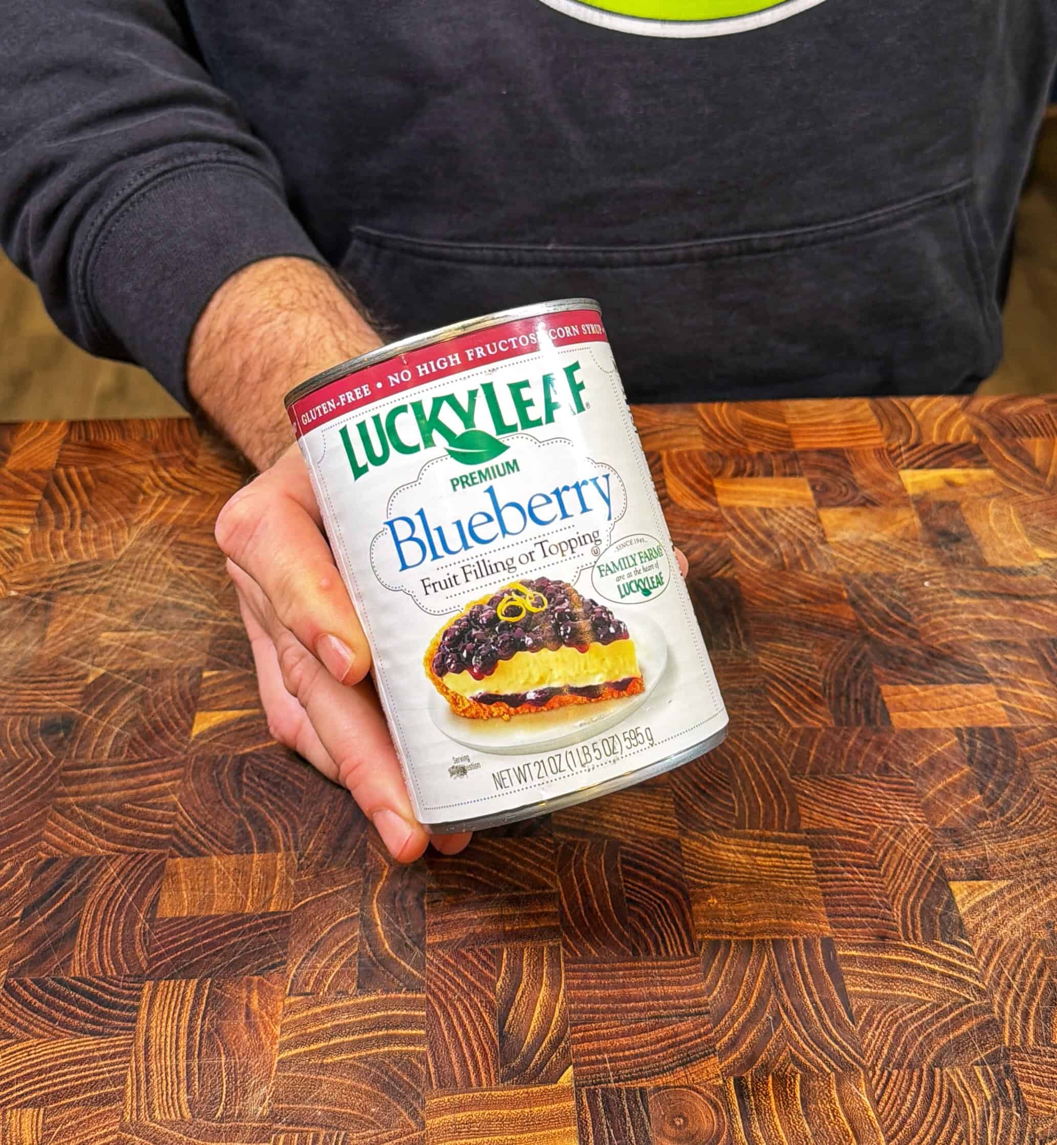 A person wearing a dark sweatshirt holds a can of Lucky Leaf Premium Blueberry Fruit Filling & Topping over a wooden countertop.