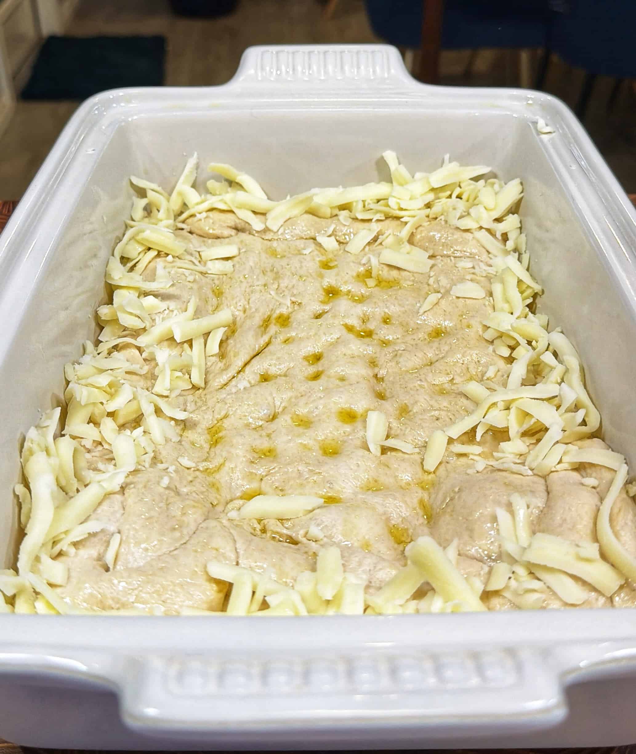 A rectangular white baking dish contains uncooked focaccia dough sprinkled with shredded cheese and drizzled with olive oil, ready to be baked.