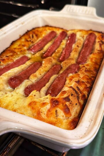 A baked dish in a white rectangular pan shows browned, fluffy batter with several hot dogs partially embedded on top, just out of the oven.