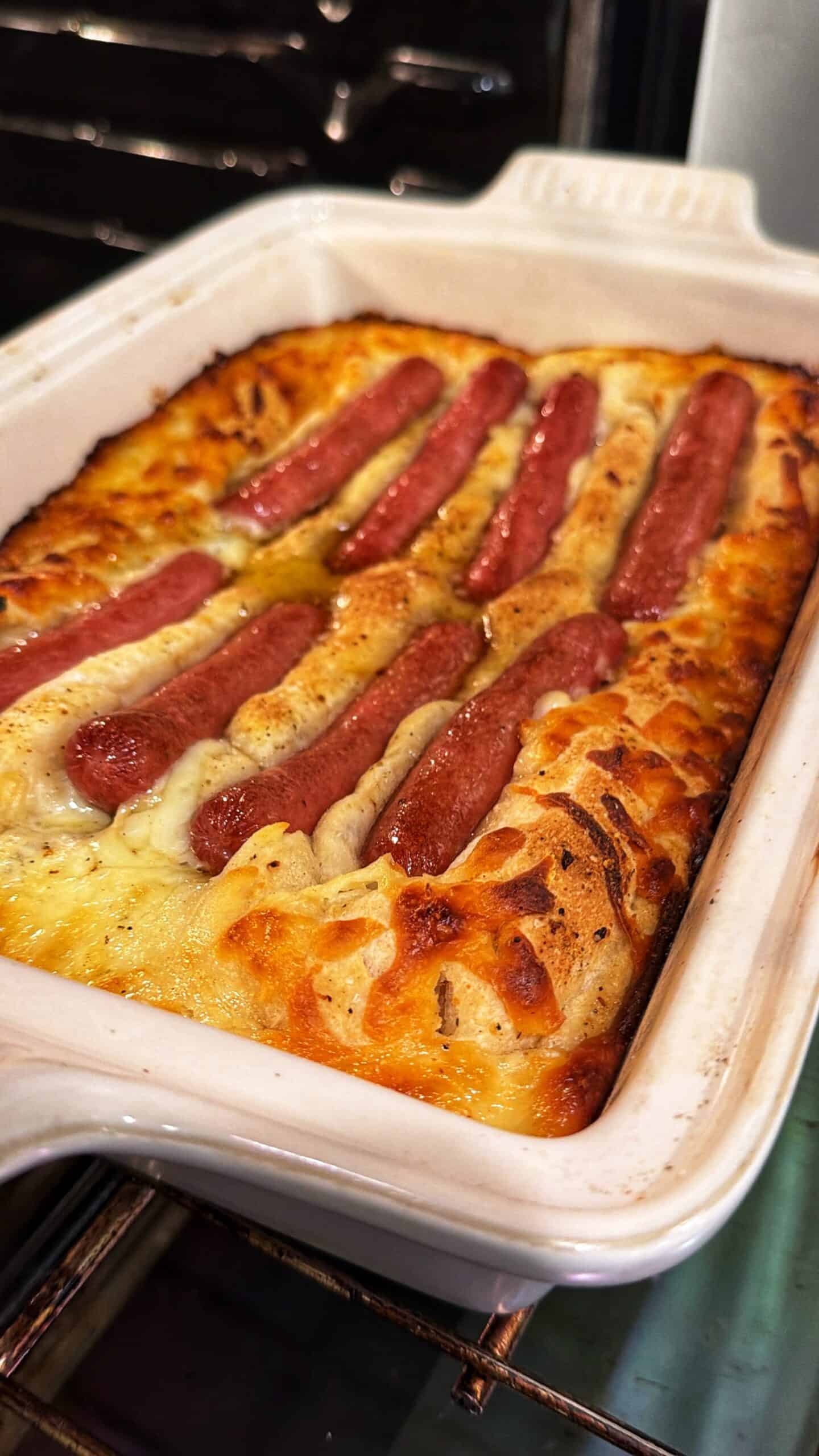 A baked dish in a white rectangular pan shows browned, fluffy batter with several hot dogs partially embedded on top, just out of the oven.